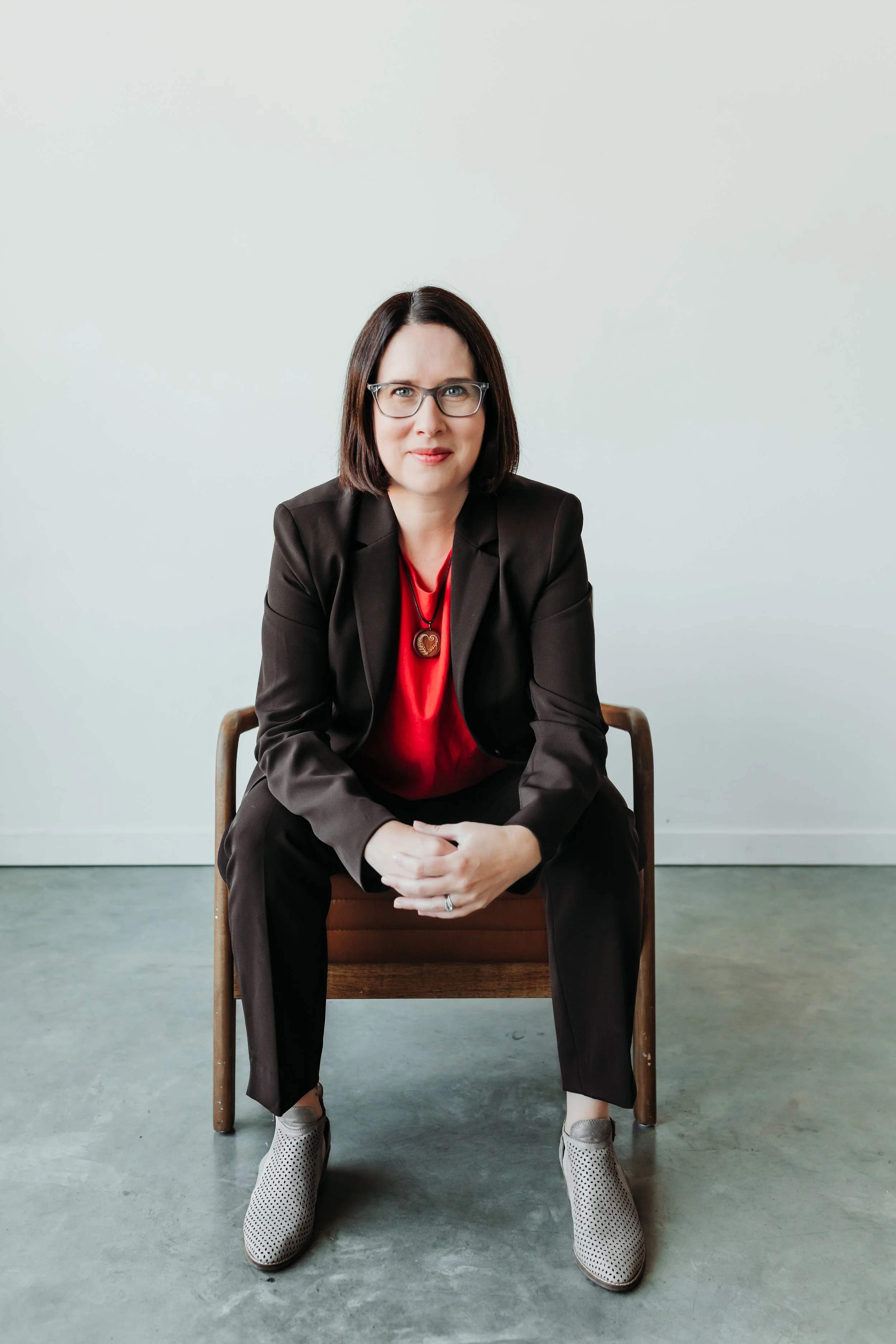 A woman with medium length brown hair, wearing glasses, a pink shirt, and a brown suit, sitting on a brown leather chair with her hands clasped, looking at the camera against a neutral background.