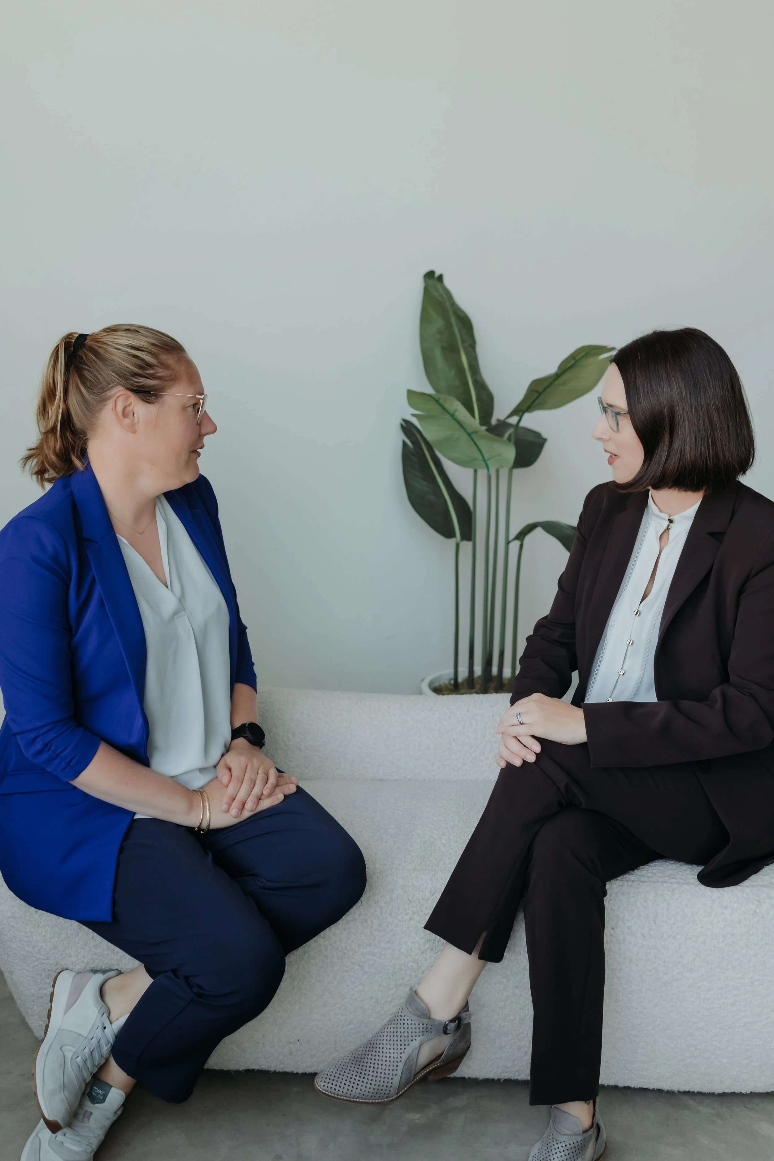 Two women sitting on a coach in conversation. The woman on the left has long blonde hair, a blue blazer and a white top. The woman on the right has medium length brown hair, a white shirt and a brown suit, against a neutral background.