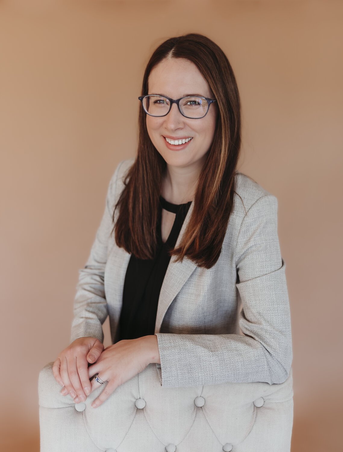 A woman with long brown hair and glasses, wearing a light-colored blazer and black top, smiling and leaning on a cream-colored upholstered chair, against a plain beige background.