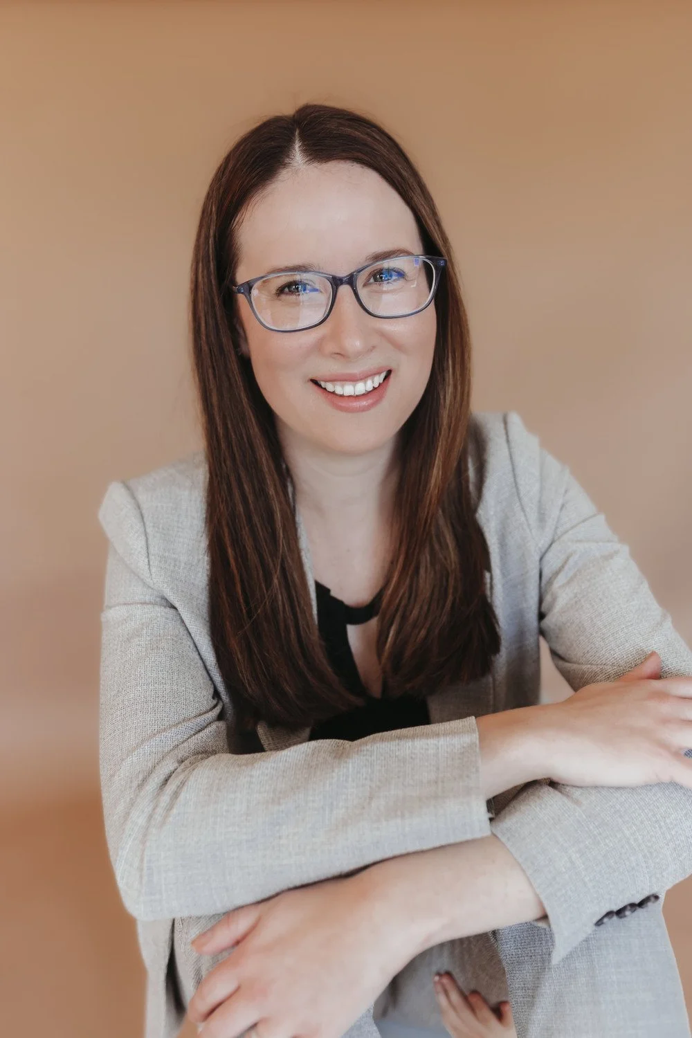 A woman with long brown hair and glasses smiling at the camera, wearing a beige blazer over a black top, sitting against a plain beige background.