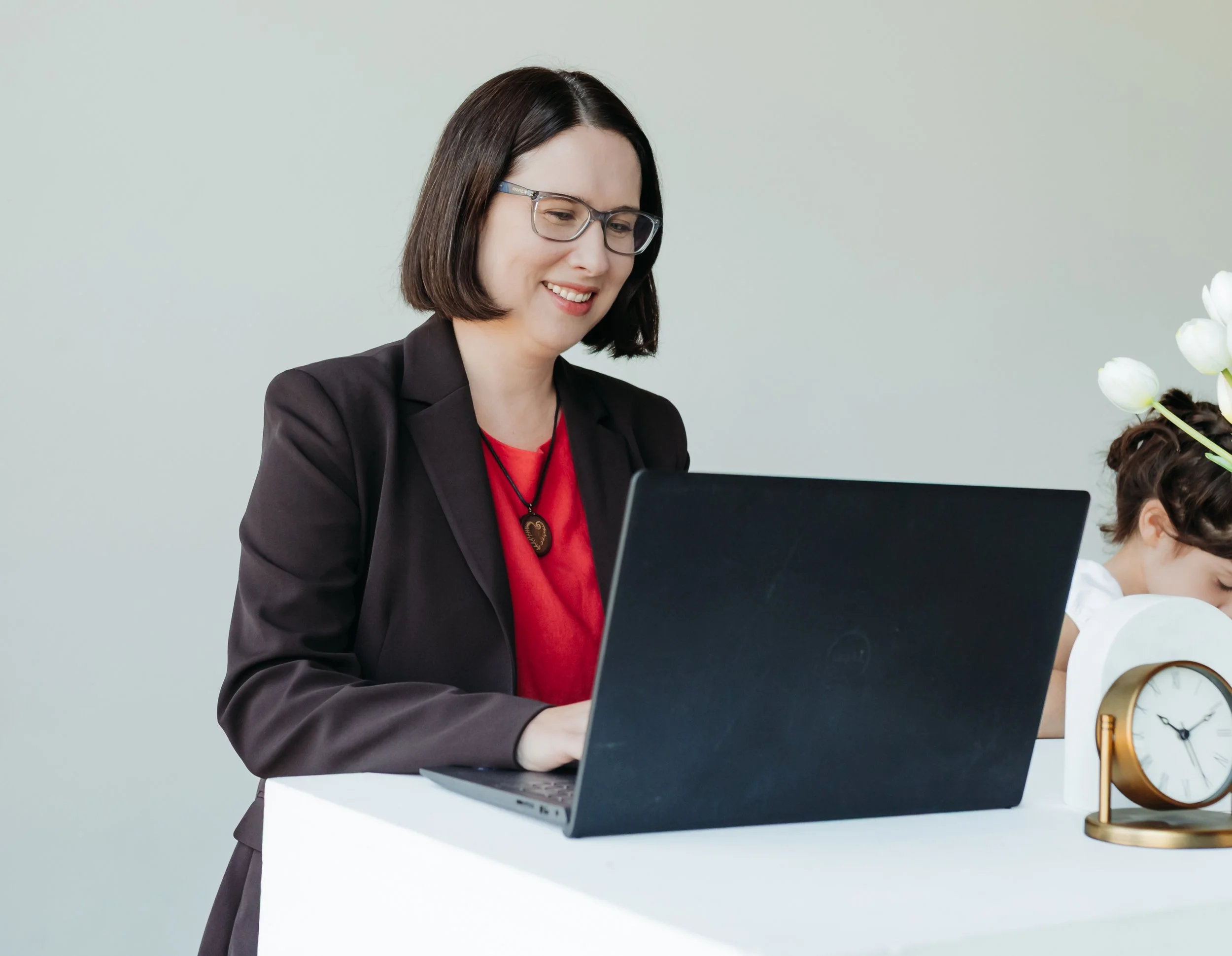 A woman with medium length brown hair and glasses in a pink shirt and brown blazer working at a laptop on a white desk beside a girl with brown hair and a clock on the desk