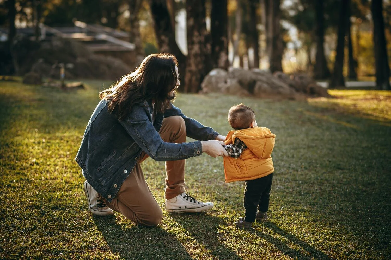 A woman kneeling on the grass holding the hands of a young boy wearing an orange vest in a park during sunset.