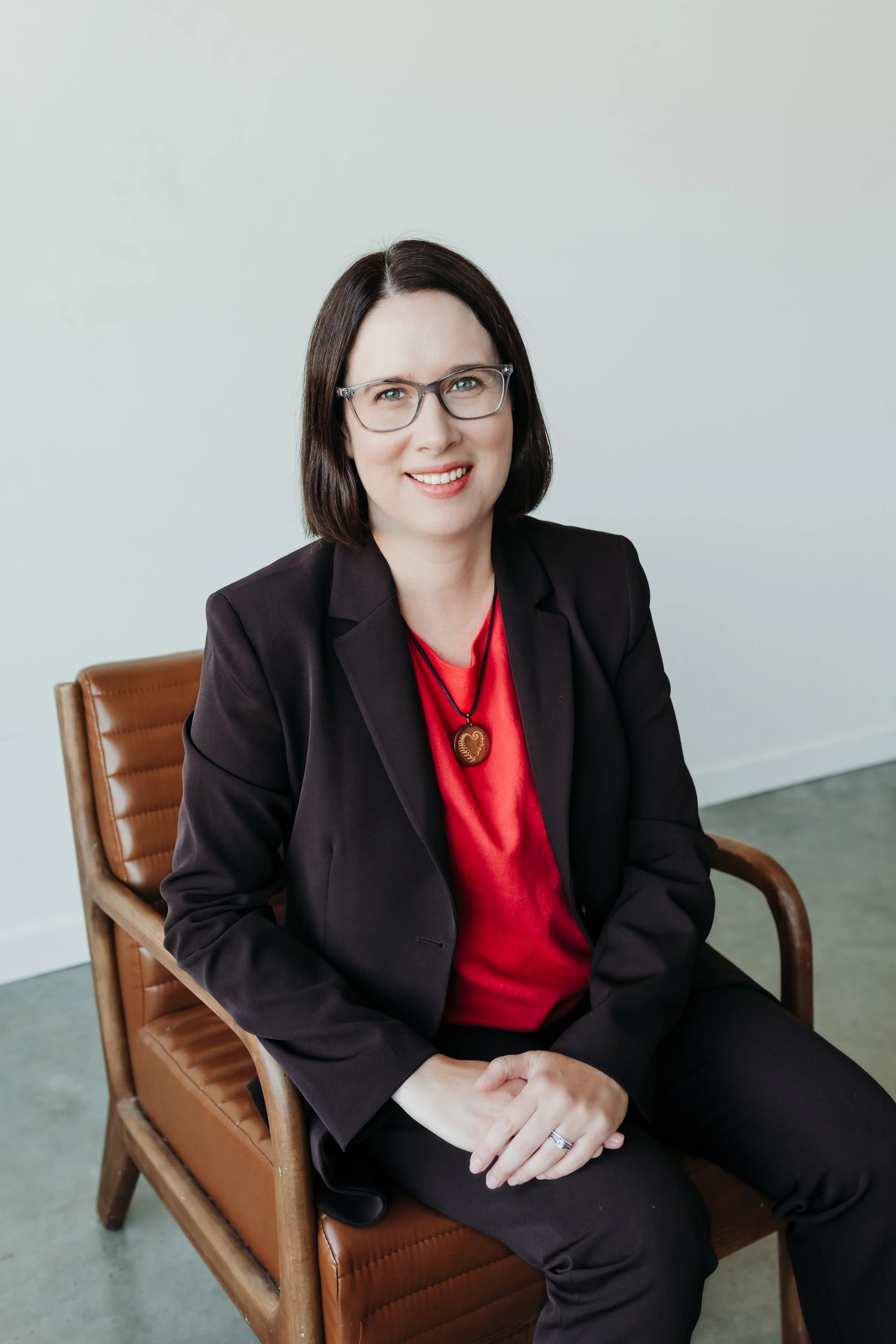 A woman with medium length brown hair, wearing glasses, a pink shirt, and a brown suit, sitting on a brown leather chair with her hands clasped, smiling at the camera against a neutral background.