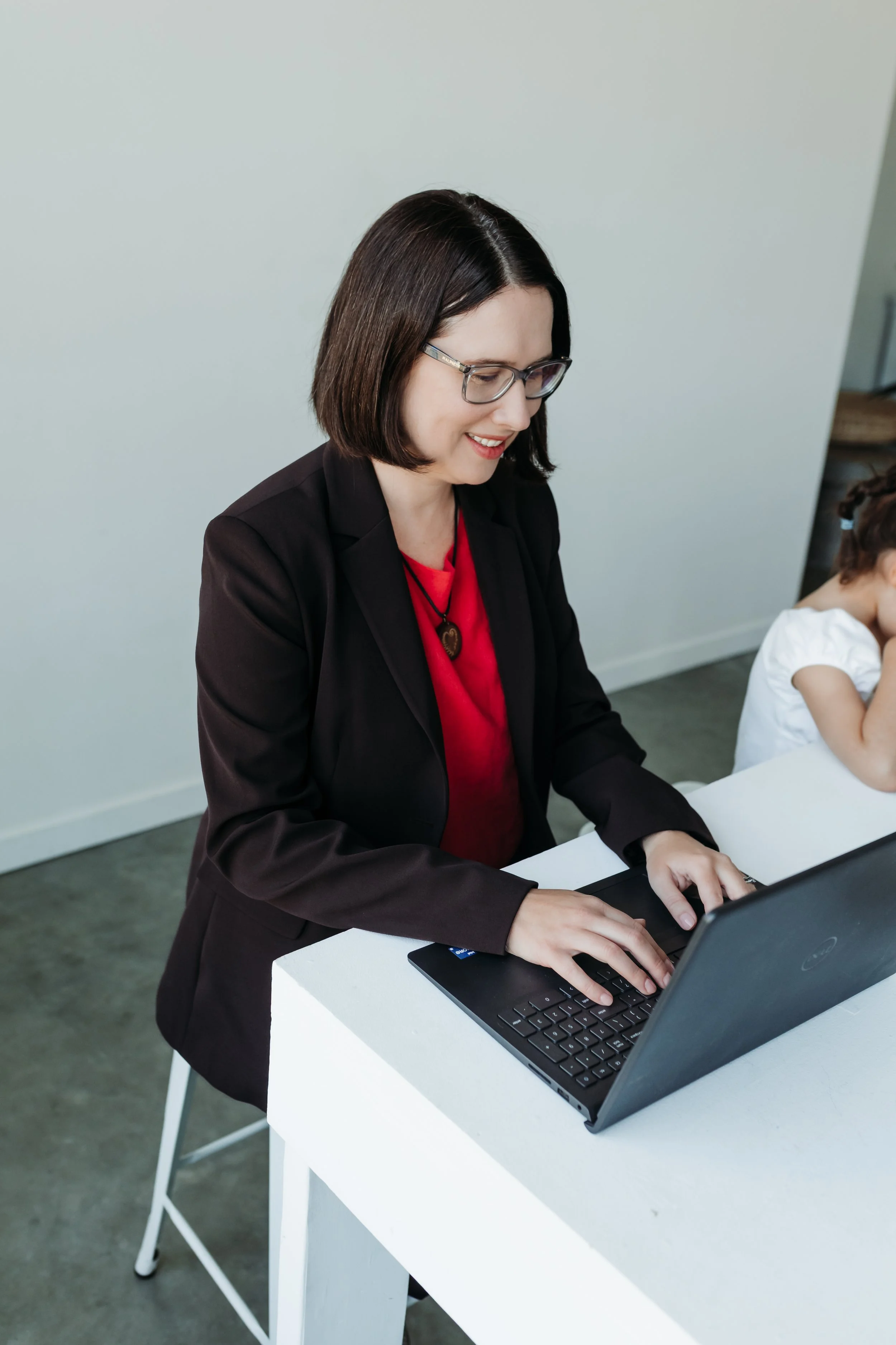 A woman with medium length brown hair and glasses wearing a pink shirt and brown blazer working at a laptop with a child sitting next to her and a neutral background