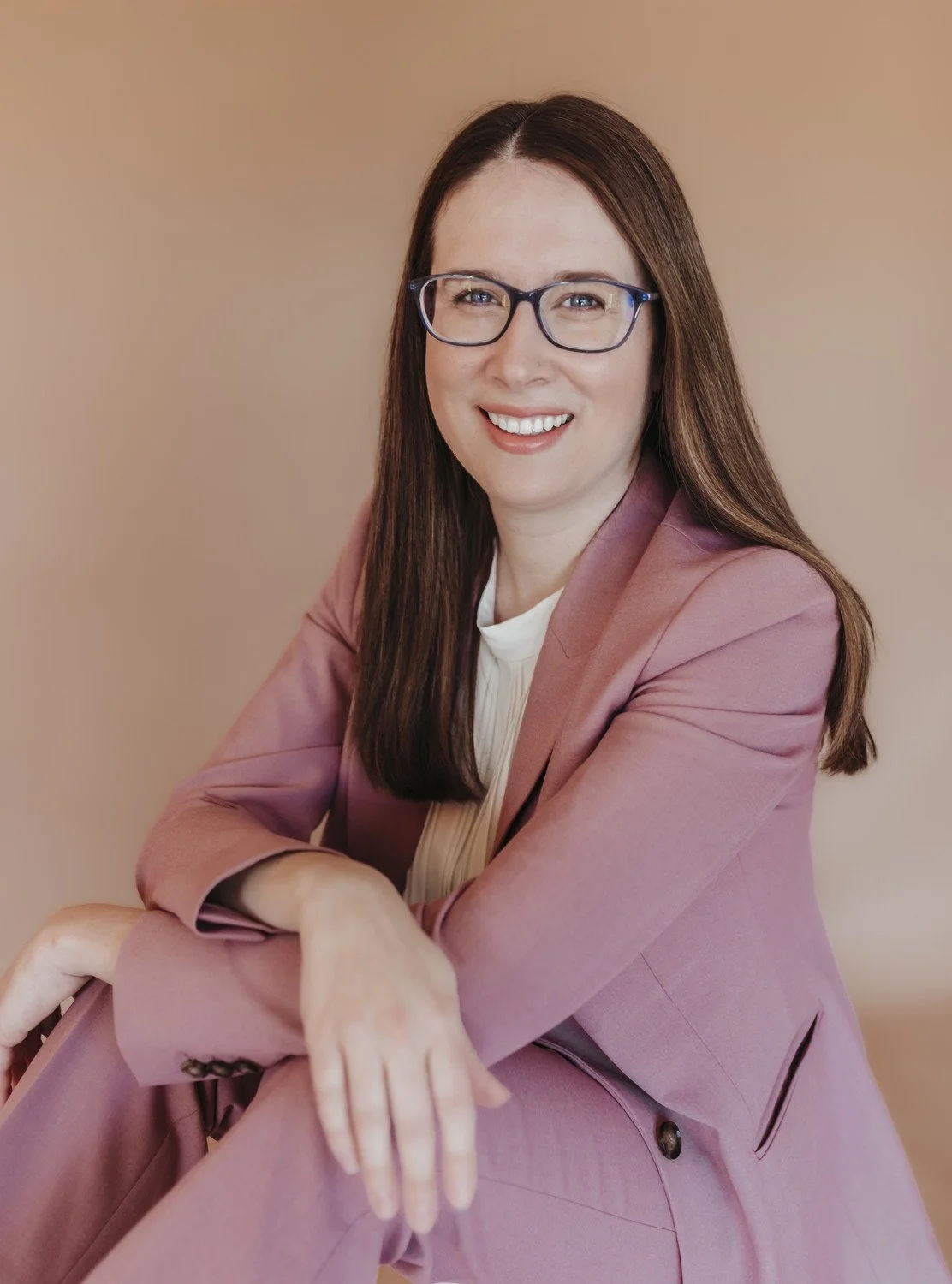 A woman with long brown hair and glasses smiling in a pink blazer and white top against a beige background.
