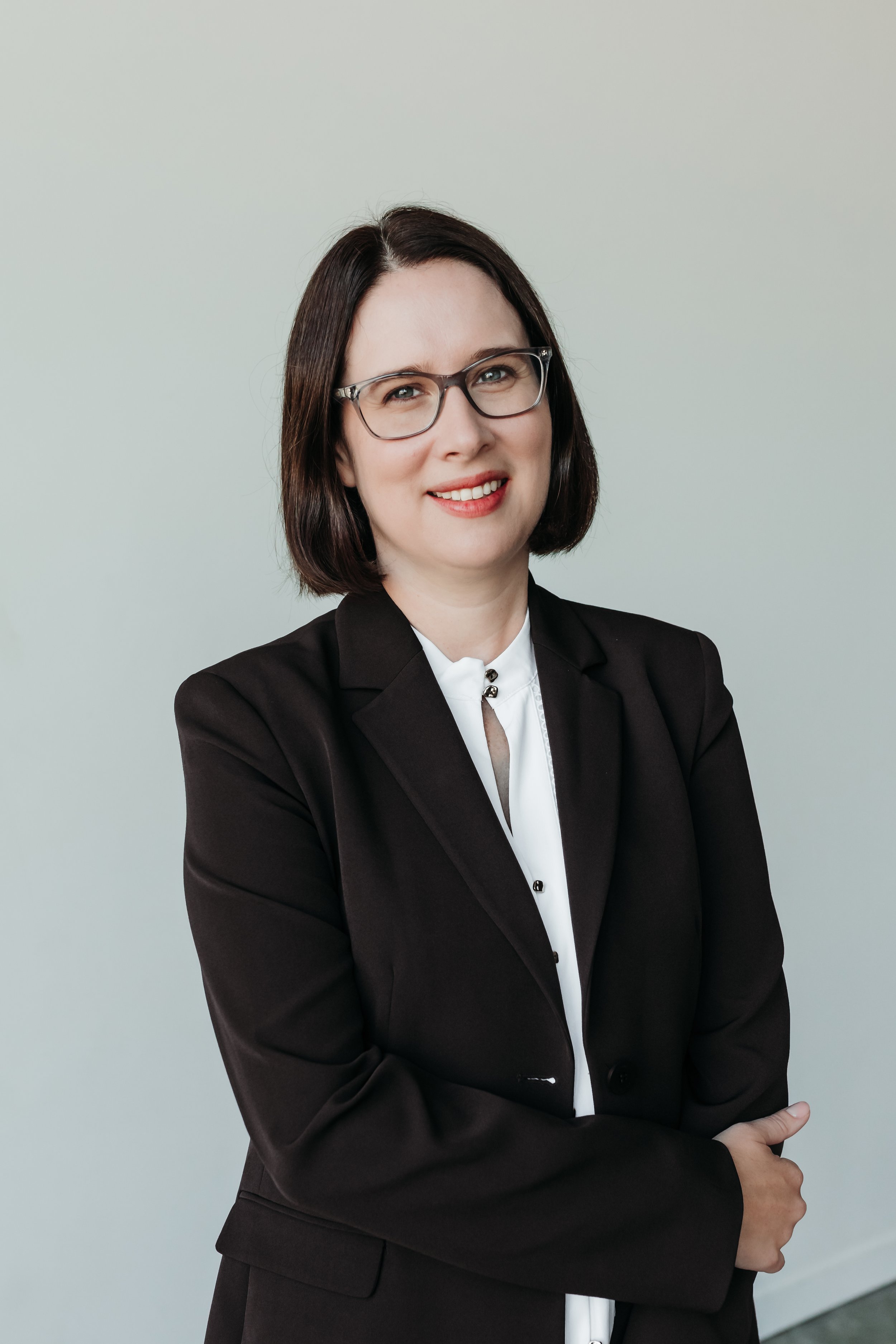 A woman with medium length brown hair and glasses wearing a white shirt and brown blazer, smiling at the camera with a neutral background.