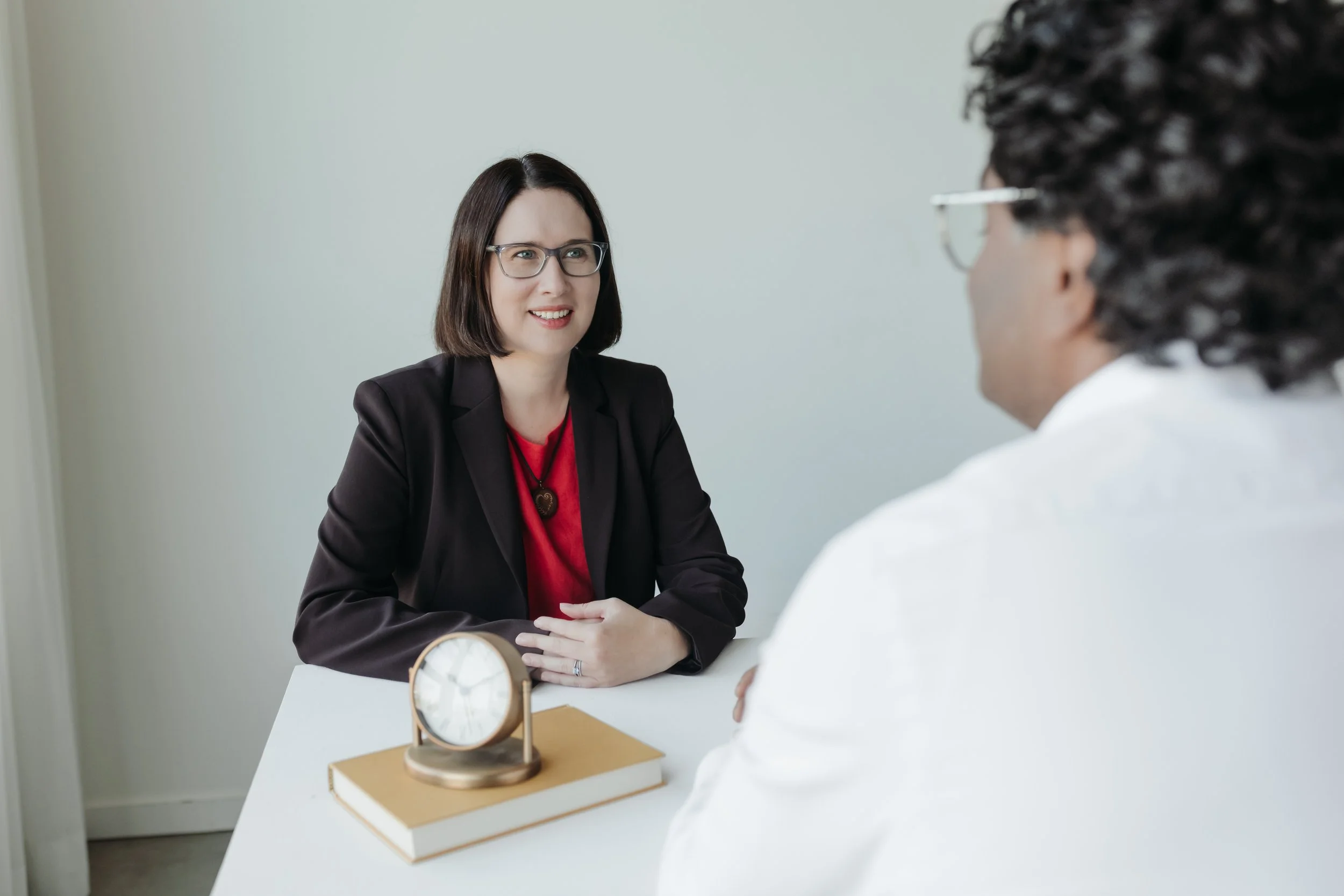 A woman with medium length brown hair, wearing glasses,brown blazer and pink top sitting at a desk in conversation with a man with glasses and curly brown hair, against a neutral background with a clock and book in the foreground.