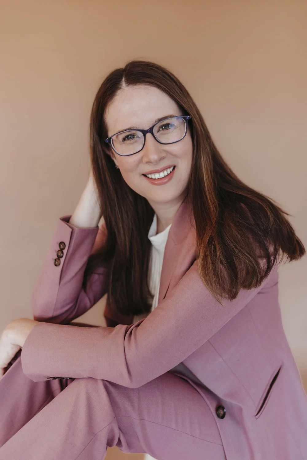 A woman with long brown hair, wearing glasses, a white shirt, and a pink blazer, sitting with her hand resting on her head, smiling at the camera against a neutral background.