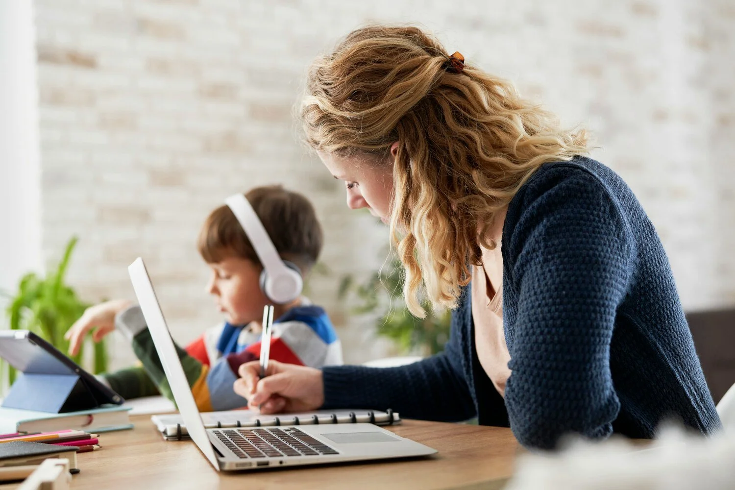 A woman helps a young boy with homework at a desk, with a laptop, a tablet, and school supplies on the table.