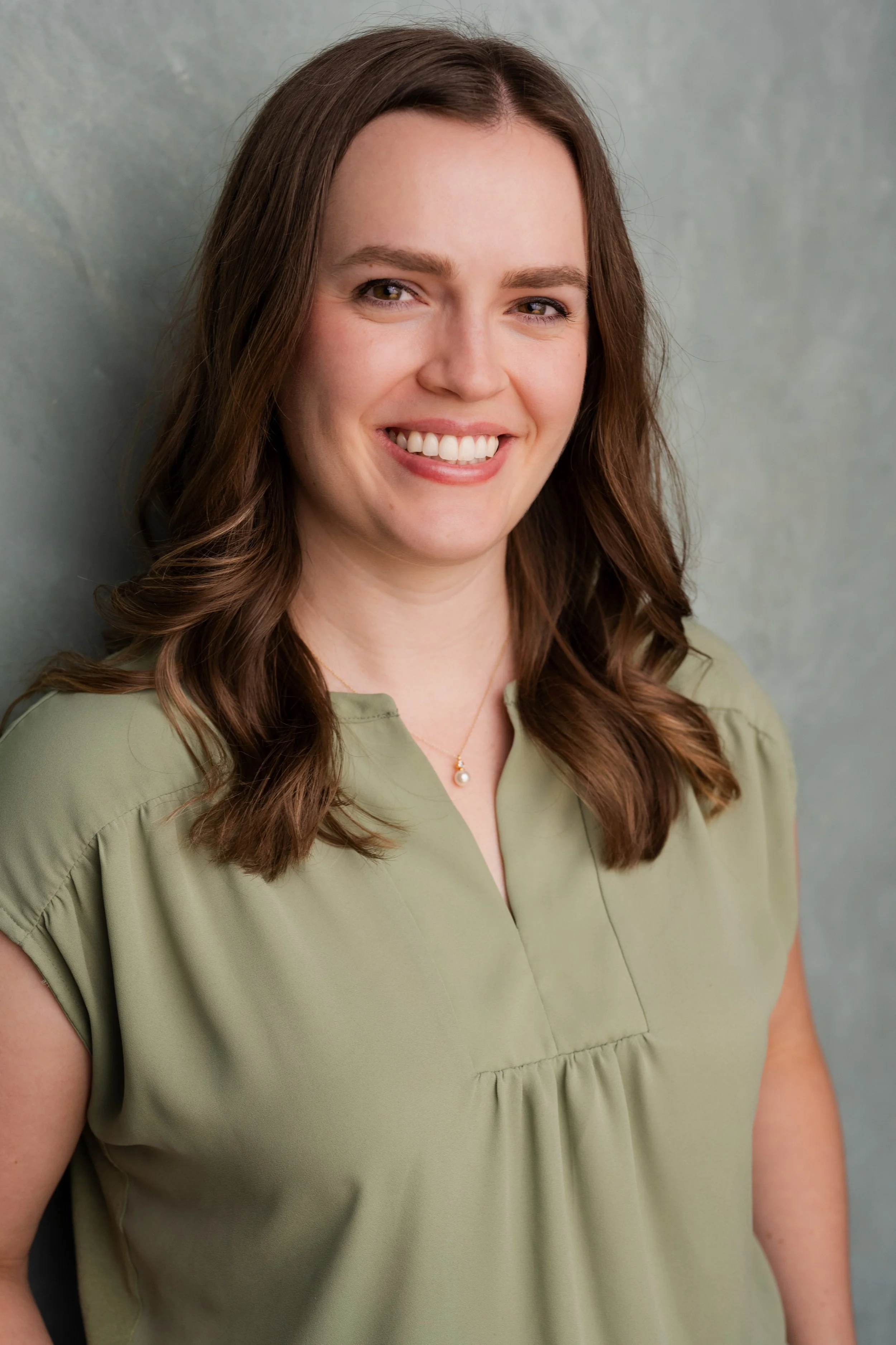 A woman with long brown hair, smiling, wearing a light green blouse and a necklace with a pearl pendant, standing against a textured green wall.