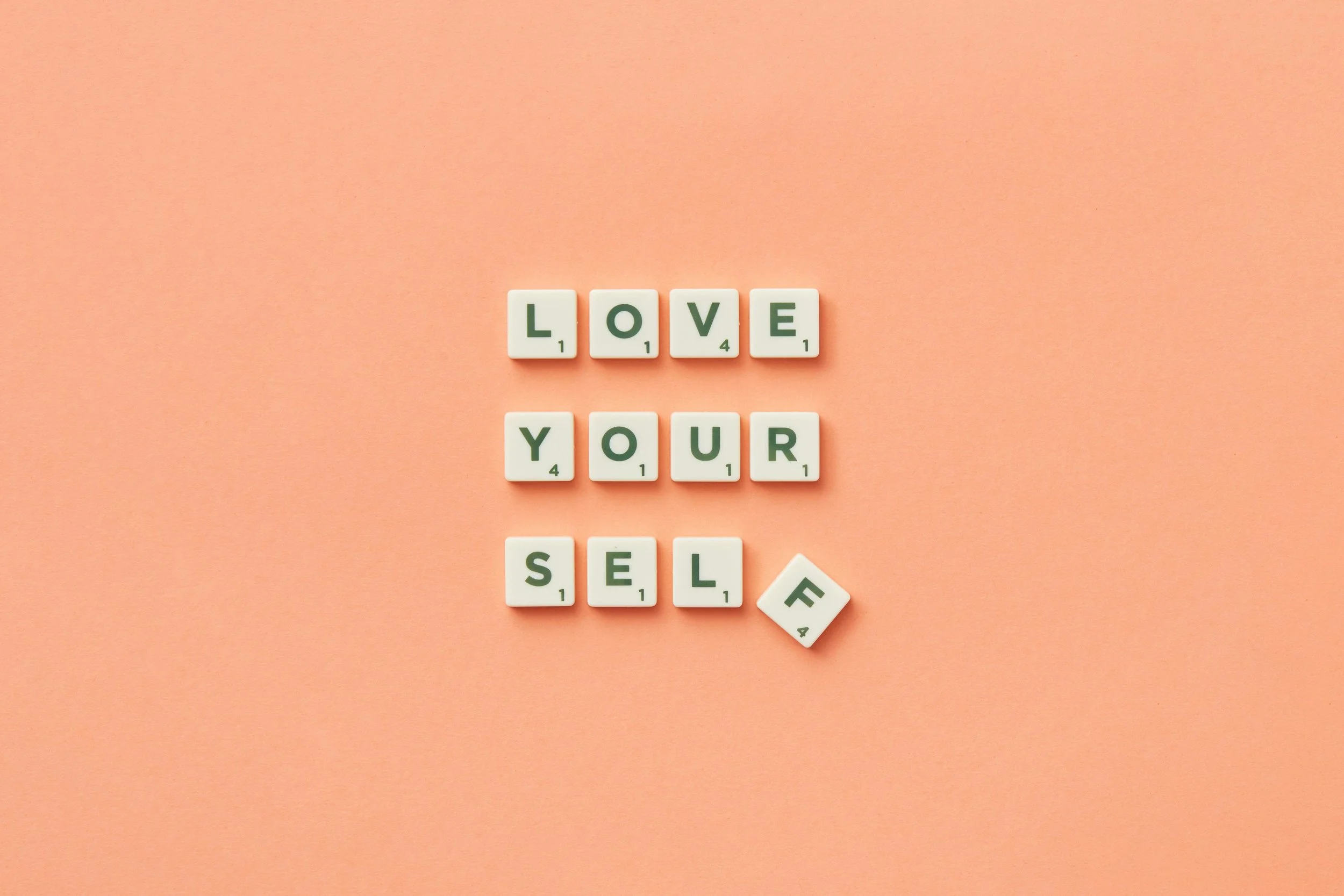 Scrabble tiles spelling out "Love Your Self" on a pink background.