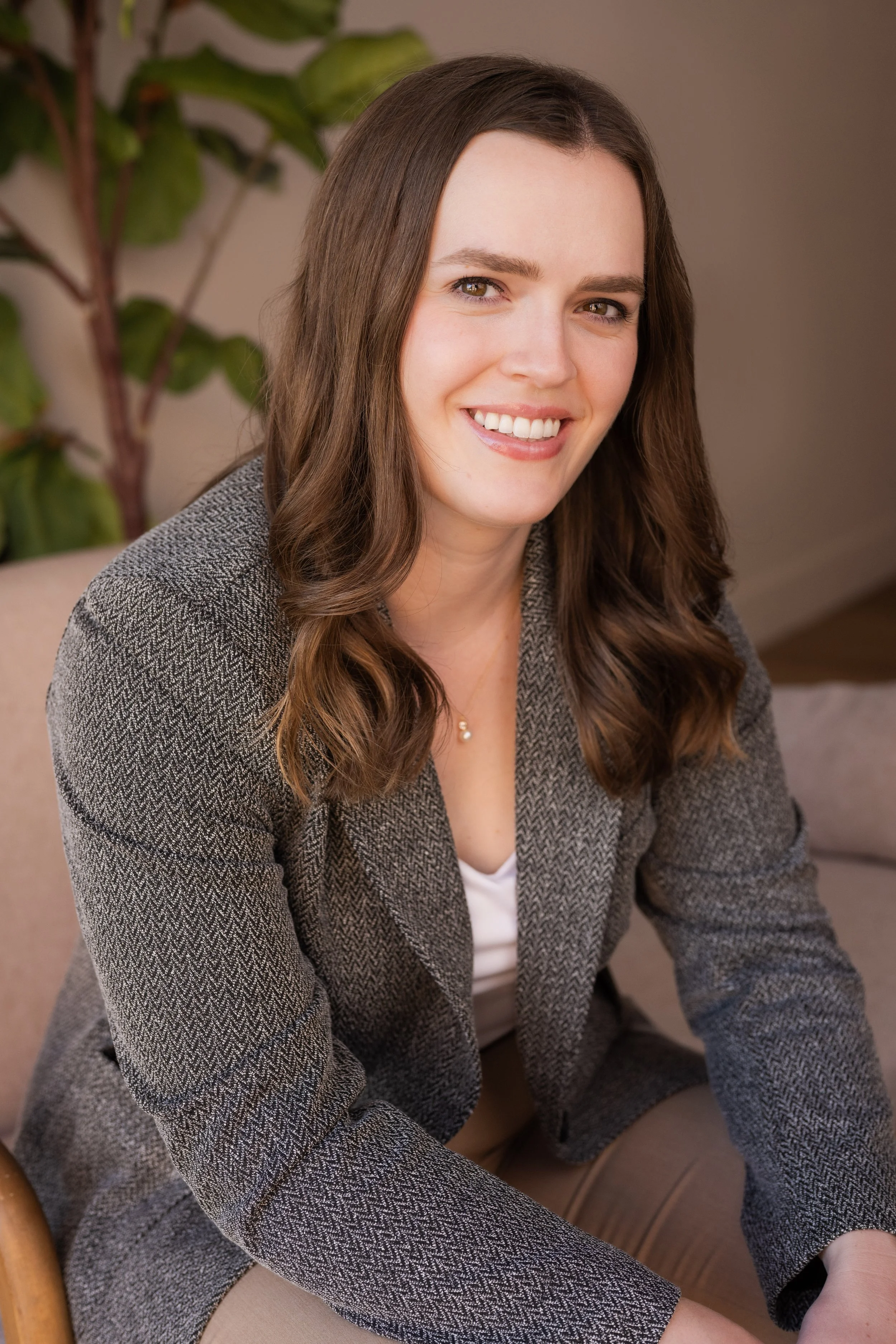 A woman with long brown hair, smiling, wearing a gray blazer and sitting on a beige chair in a room with a plant in the background.