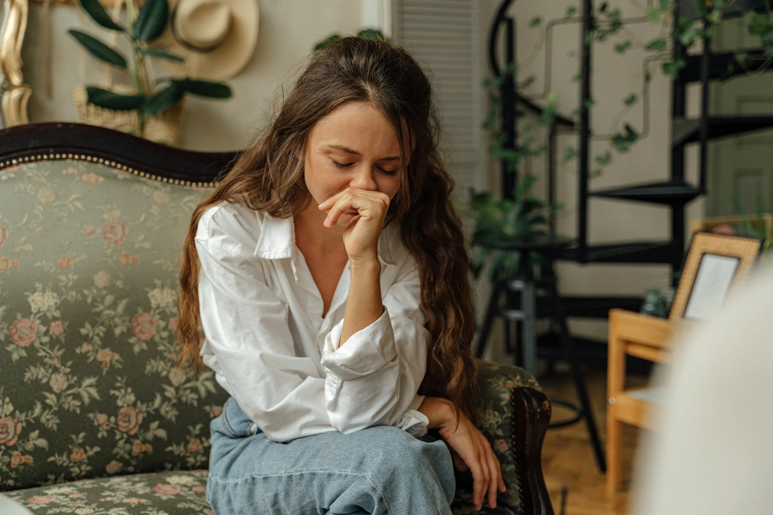 A woman with long curly hair in a white shirt sitting on a floral couch, appearing upset with her hand covering her mouth.