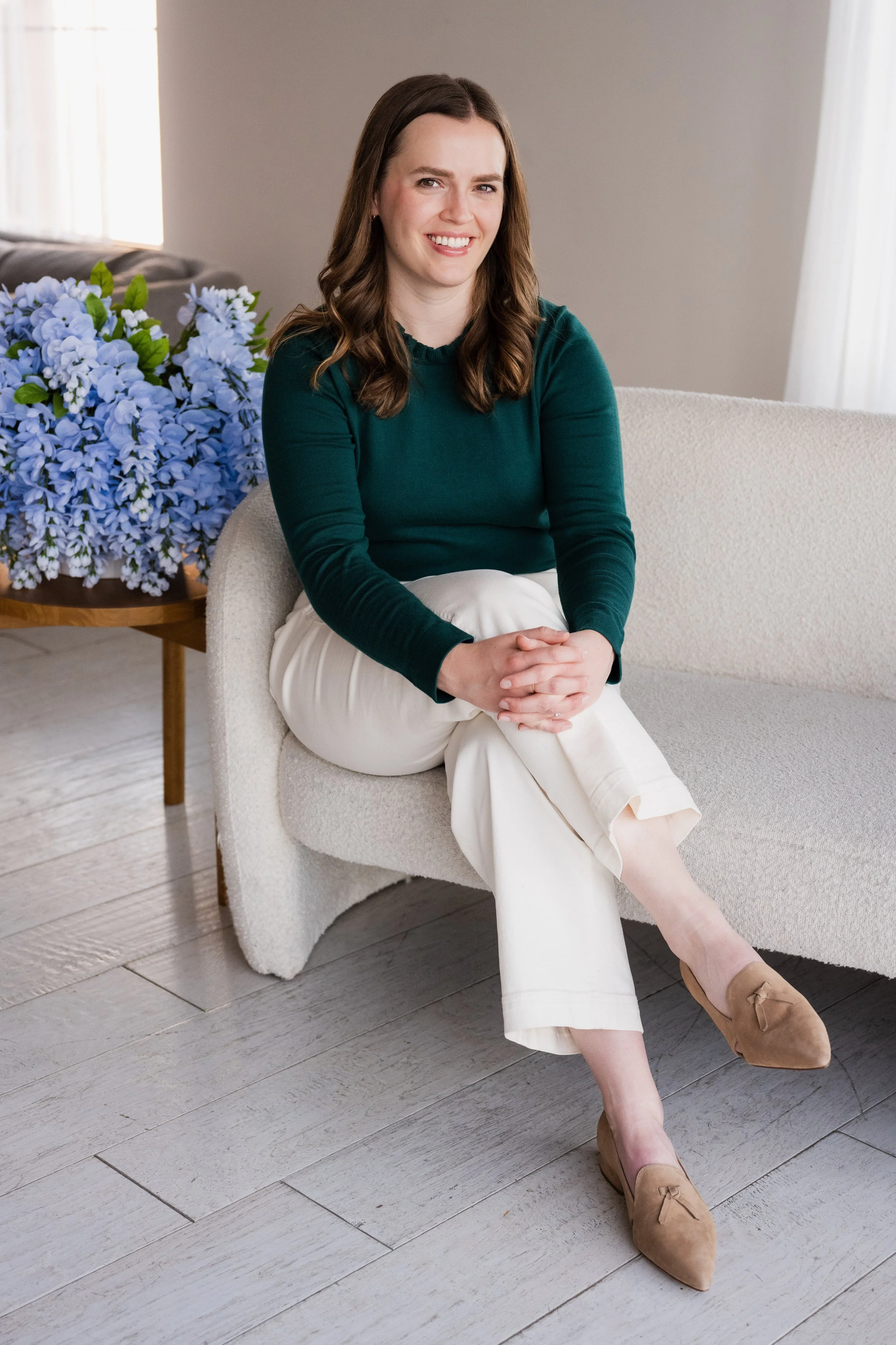 A woman with shoulder-length brown hair sitting on a white textured sofa, wearing a dark green long-sleeve top, white pants, and tan shoes, smiling with hands clasped on her lap, in a softly lit room with a window and a bunch of blue flowers on a small wooden table nearby.
