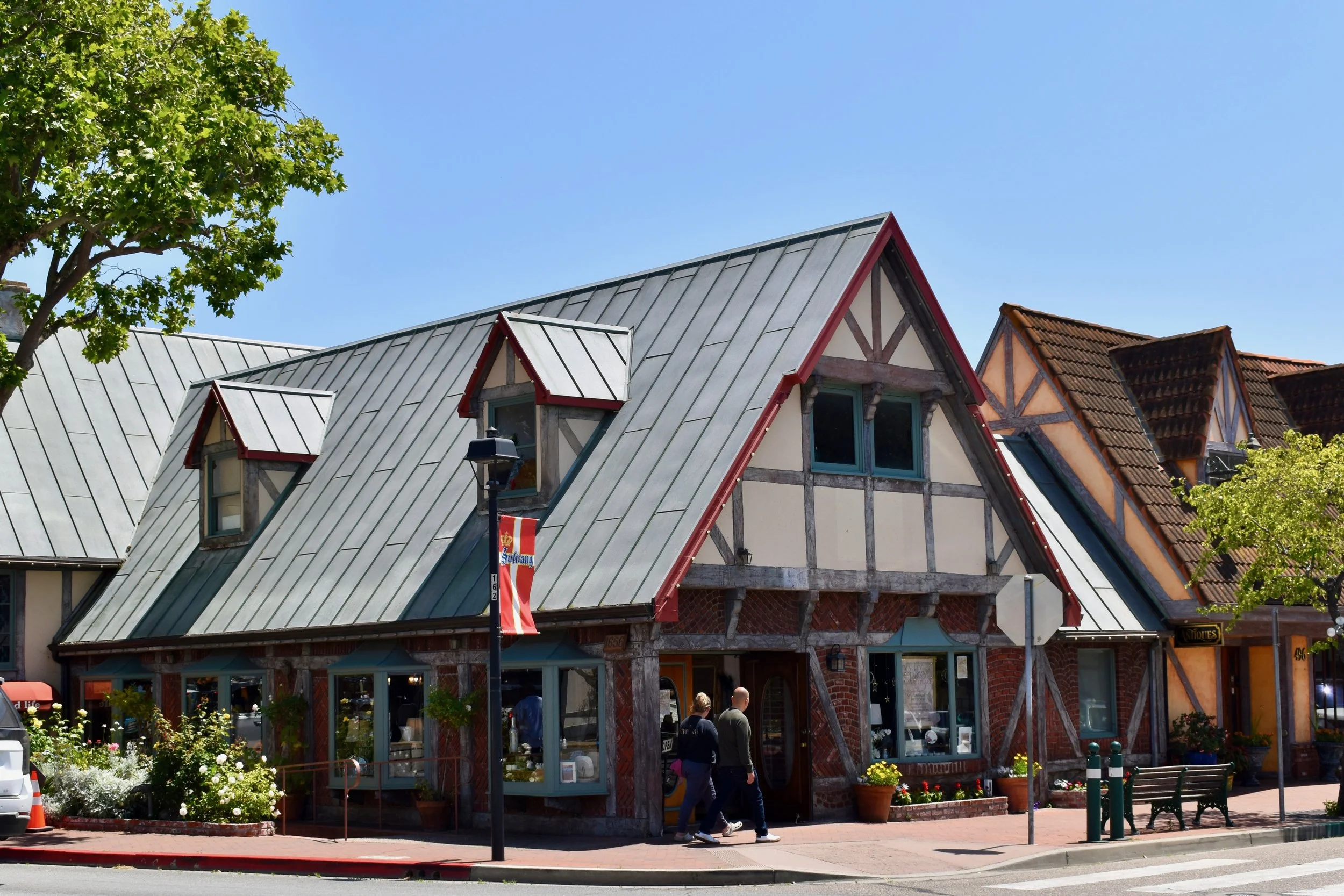 A street view of Tudor-style buildings with steep, metal roofs, dormer windows, and exposed wooden beams. There is a tree with green leaves on the left and a lamppost with a red and white banner. People are walking along the sidewalk in front of the buildings, which appear to be shops or cafes.