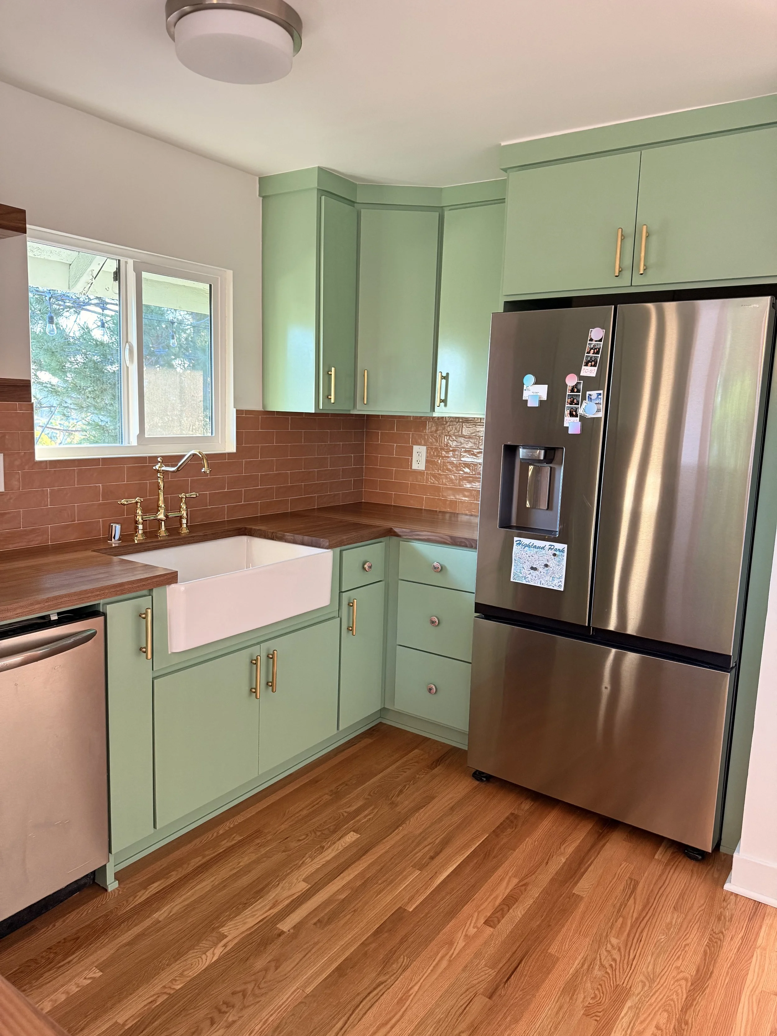 Completed kitchen corner with custom green cabinets, farmhouse sink, brass faucet, wood countertops, and stainless steel refrigerator.