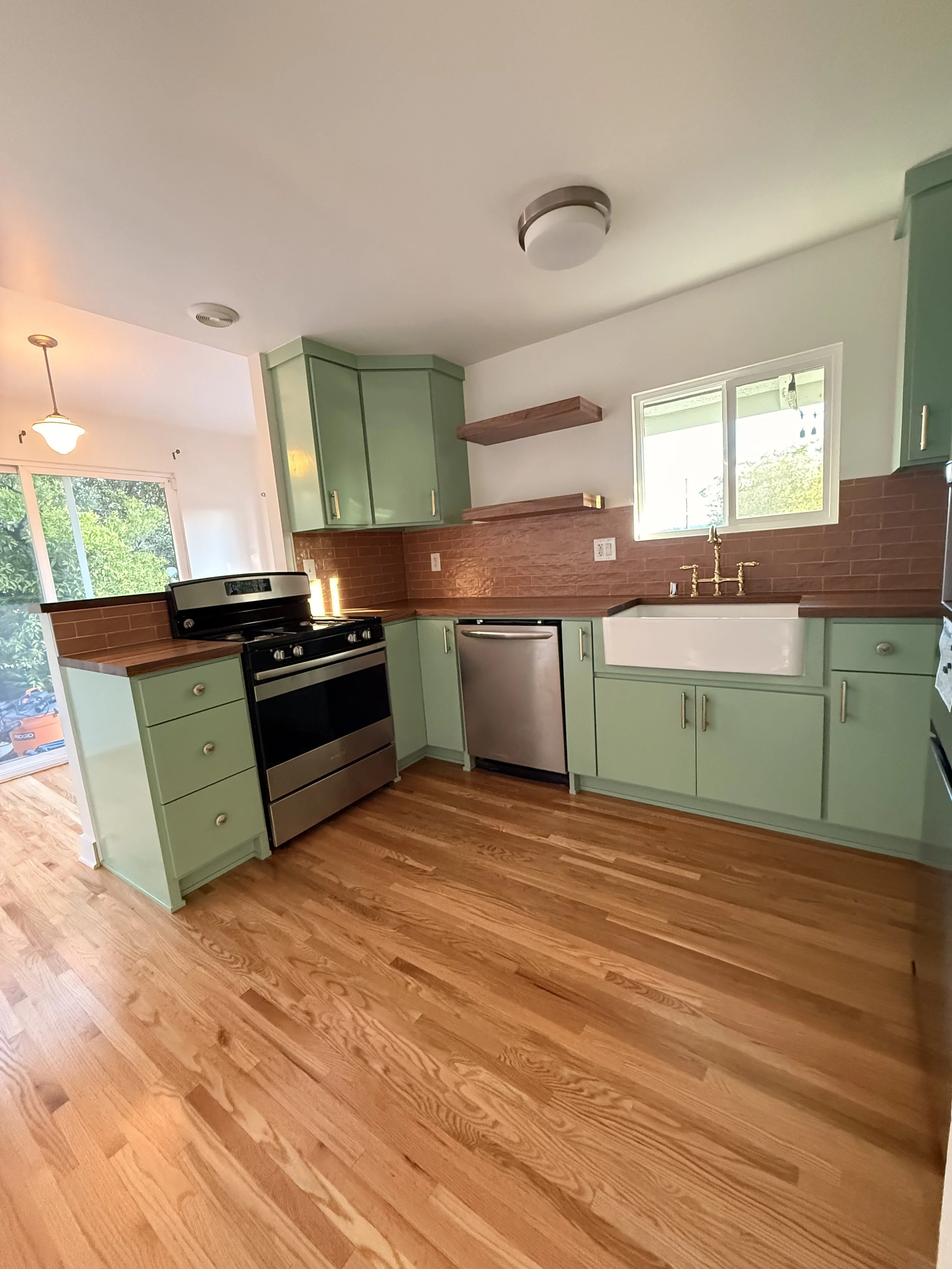 Wide view of completed kitchen remodel with green cabinetry, open wood shelving, stainless steel appliances, and warm wood floors.