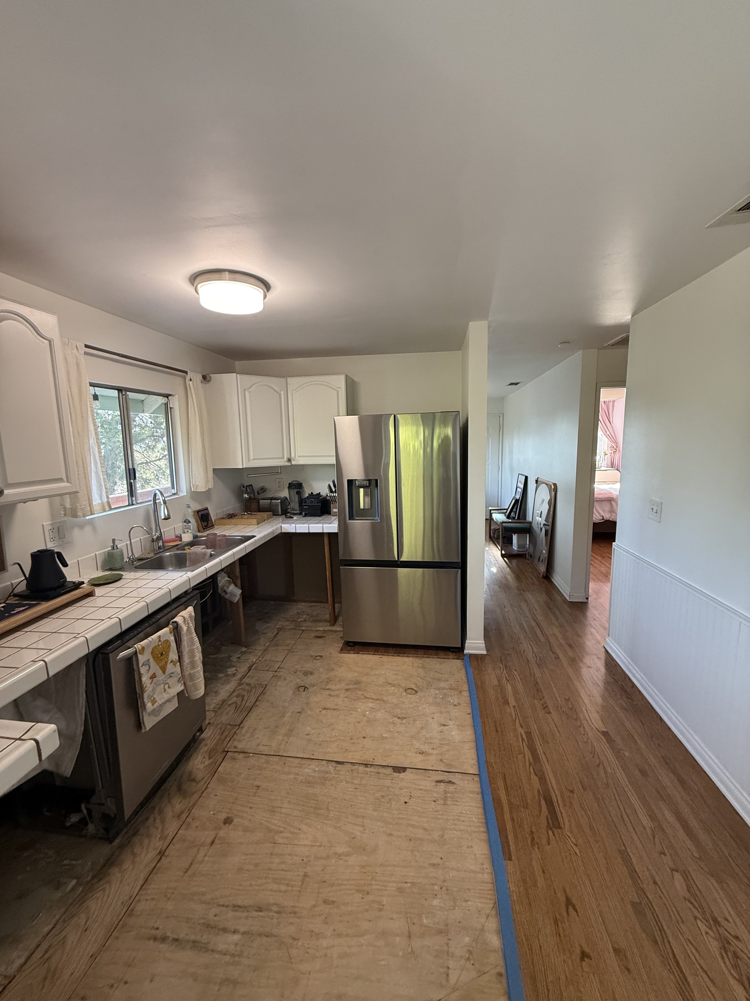 Before kitchen remodel viewed from the side, showing exposed subfloor, white tile counters, existing sink area, and stainless steel refrigerator.