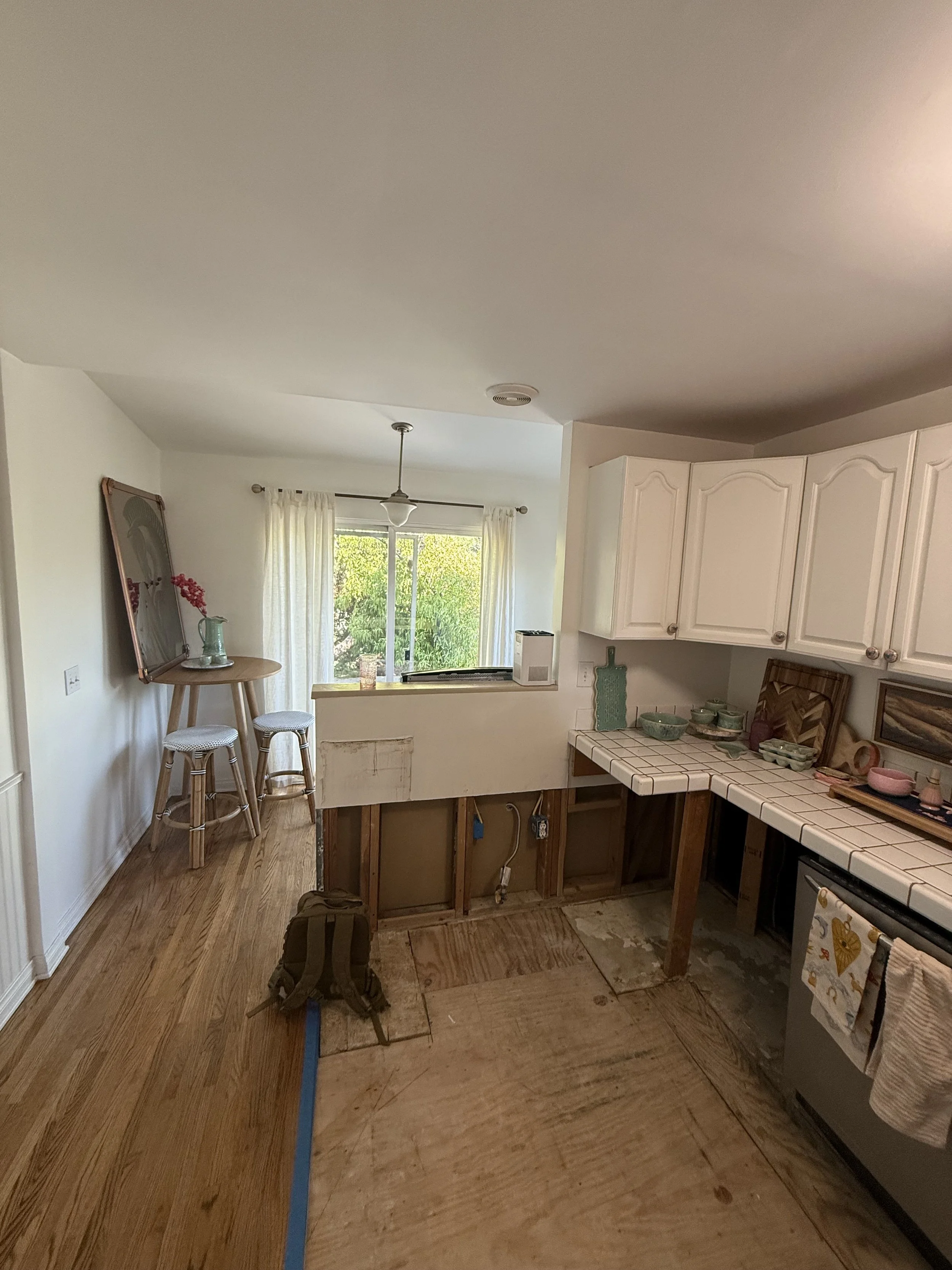 Before remodel view of kitchen and dining area with exposed wall framing below the peninsula, tile countertops, and unfinished flooring.
