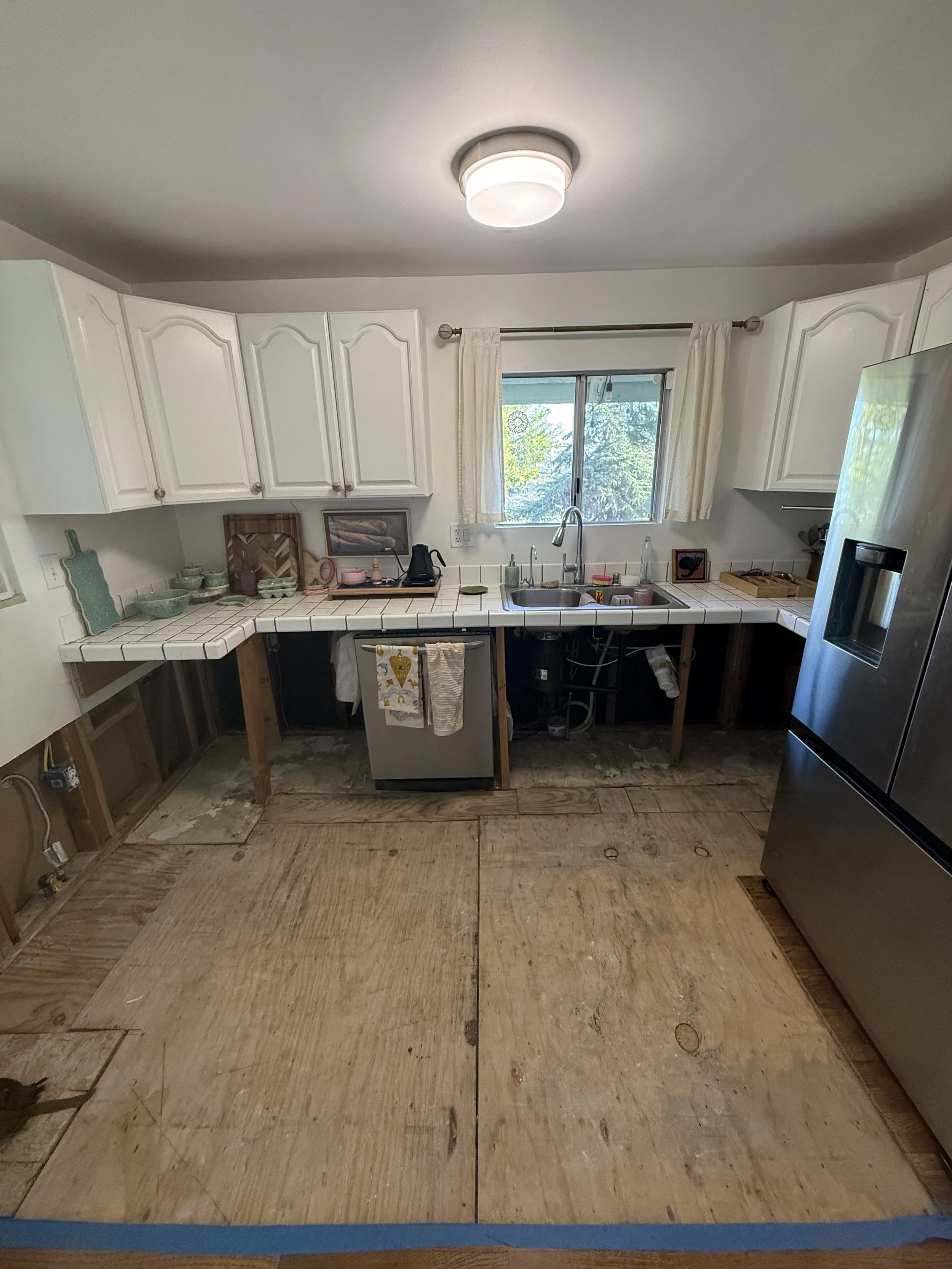 Before remodel kitchen with exposed subfloor, partially removed lower cabinetry, white upper cabinets, tile countertops, and stainless steel refrigerator.
