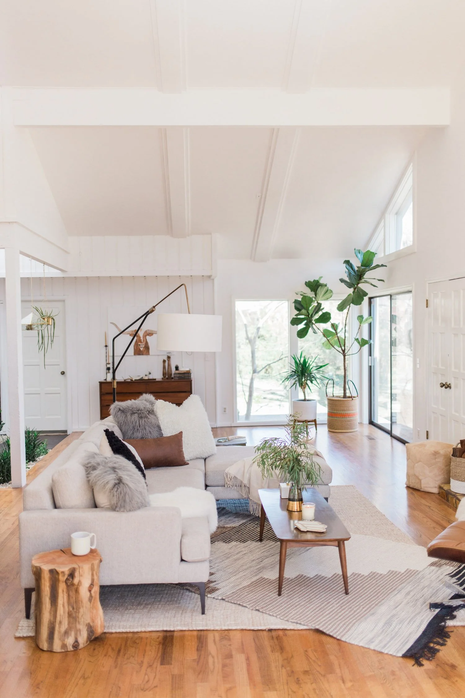 Bright living room with white walls and ceiling, large windows, beige sofa with furry pillows, wooden coffee table with plants, and various indoor plants in pots, along with a wooden side table and cozy decor.
