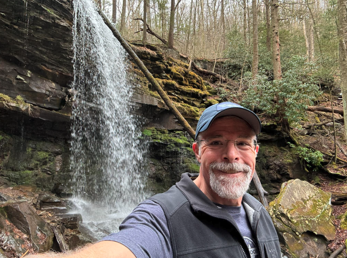 Mort Neely wearing a hat and glasses taking a selfie in front of a waterfall in the beautiful woods of rural Pennsylvania.