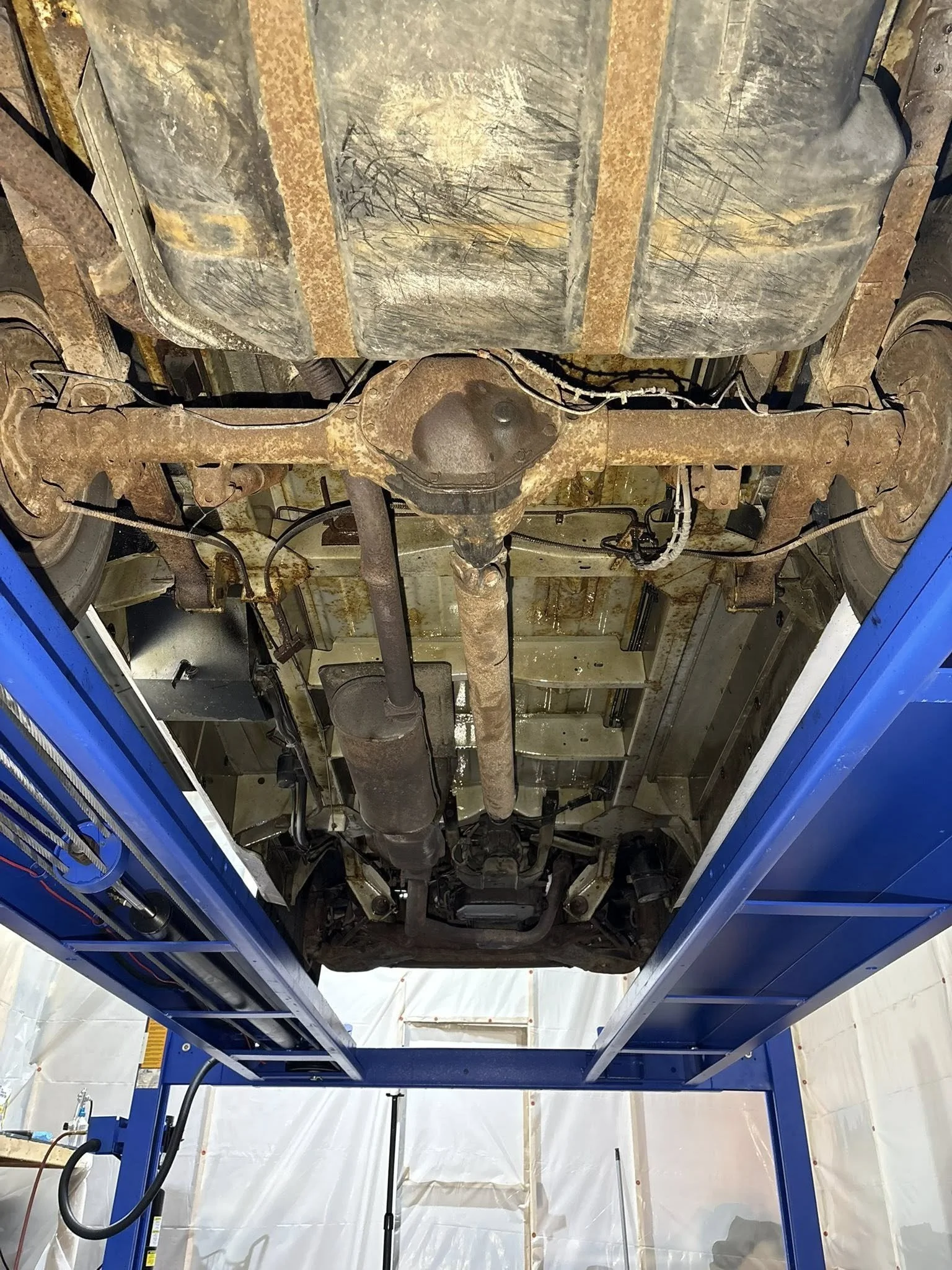 Underneath view of a vehicle on a blue lift inside IRONCLAD Vehicle Undercoating's indoor workshop showing rusted exhaust system and differential, with some rust and dirt on the underside.