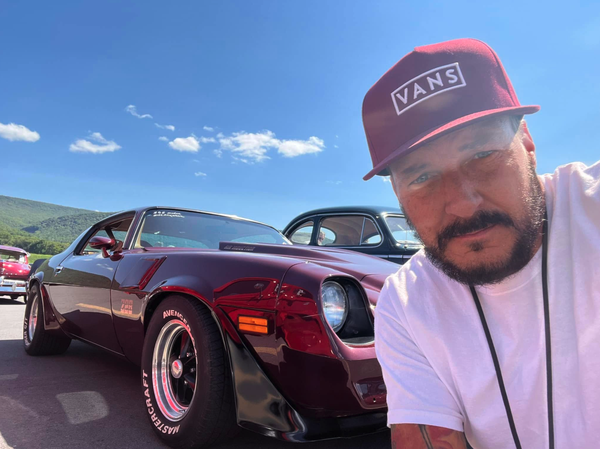 Yancy Cupp, of Cupp's Vacuum Center, ACTION Construction, and IRONCLAD Vehicle Undercoating, taking a selfie in front of a shiny red classic car with a black car behind it, under a bright blue sky with a few clouds, in a scenic outdoor setting.