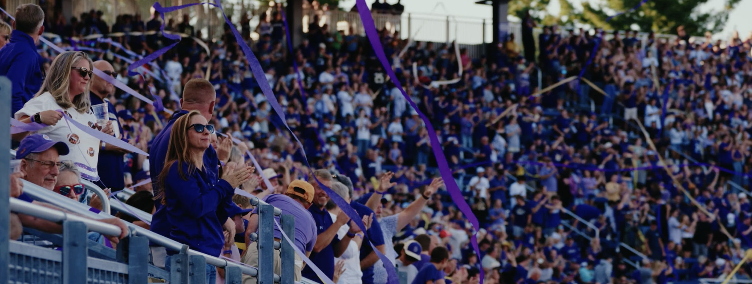 A large crowd of sports fans at a stadium, many wearing purple and clapping, with purple streamers in the air.