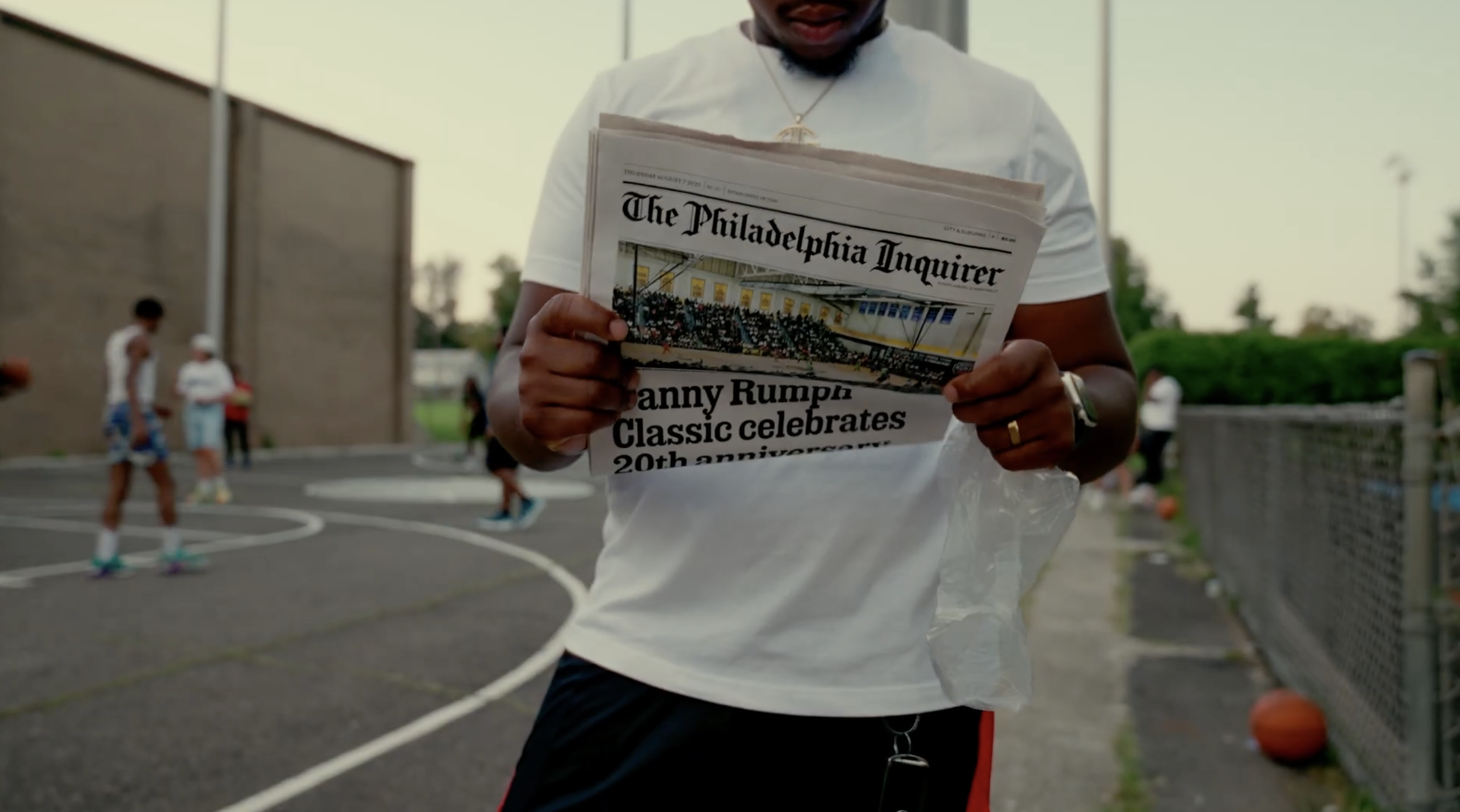 Person holding a newspaper titled 'The Philadelphia Inquirer' with a headline about a classic basketball event, standing near a basketball court with kids playing in the background.