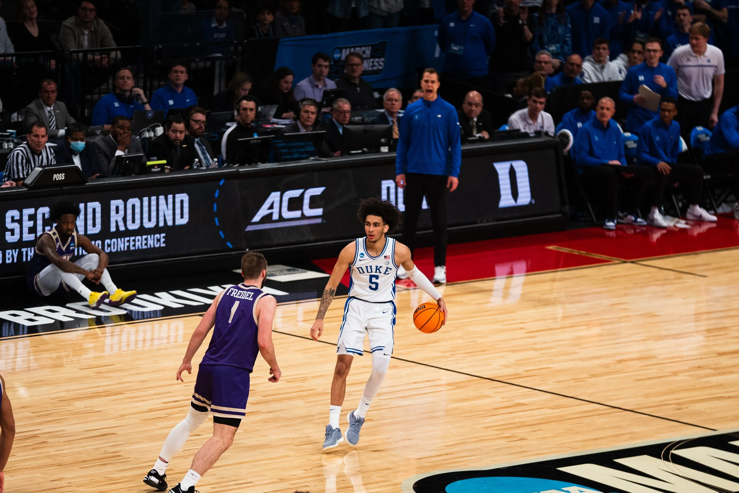 College basketball game with Duke player (number 5) dribbling the ball and an opposing player (number 1) defending. Bench and audience in the background.