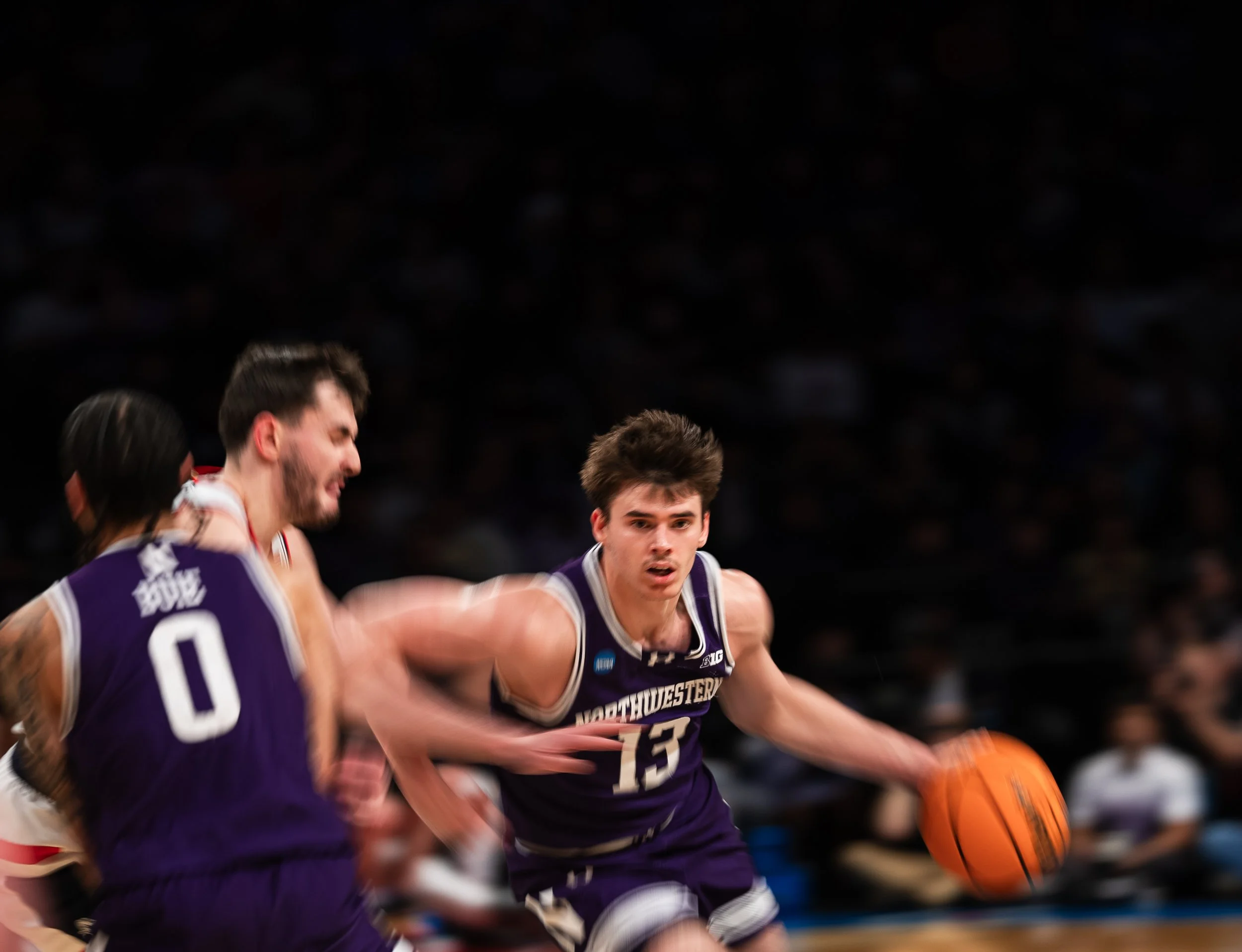 College basketball game with players competing, one in purple jersey dribbling the ball while other players defend.