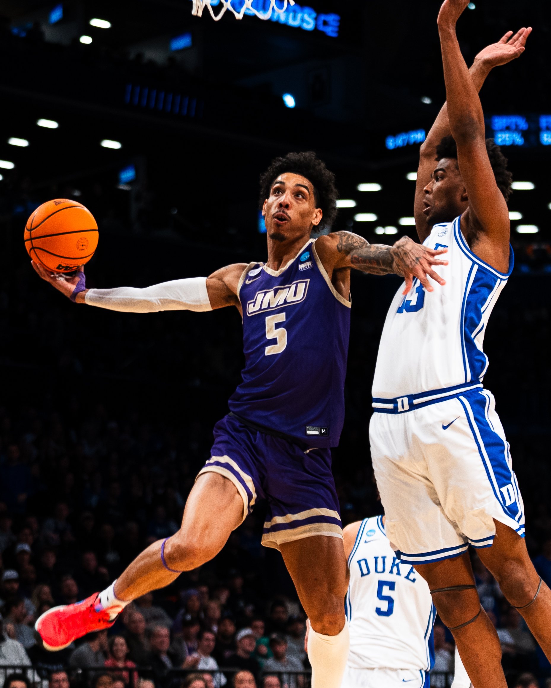 Two basketball players are mid-air during a game; one from JMU wearing a purple uniform with number 5 is attempting a shot, while the other from Duke is defending with arms raised.