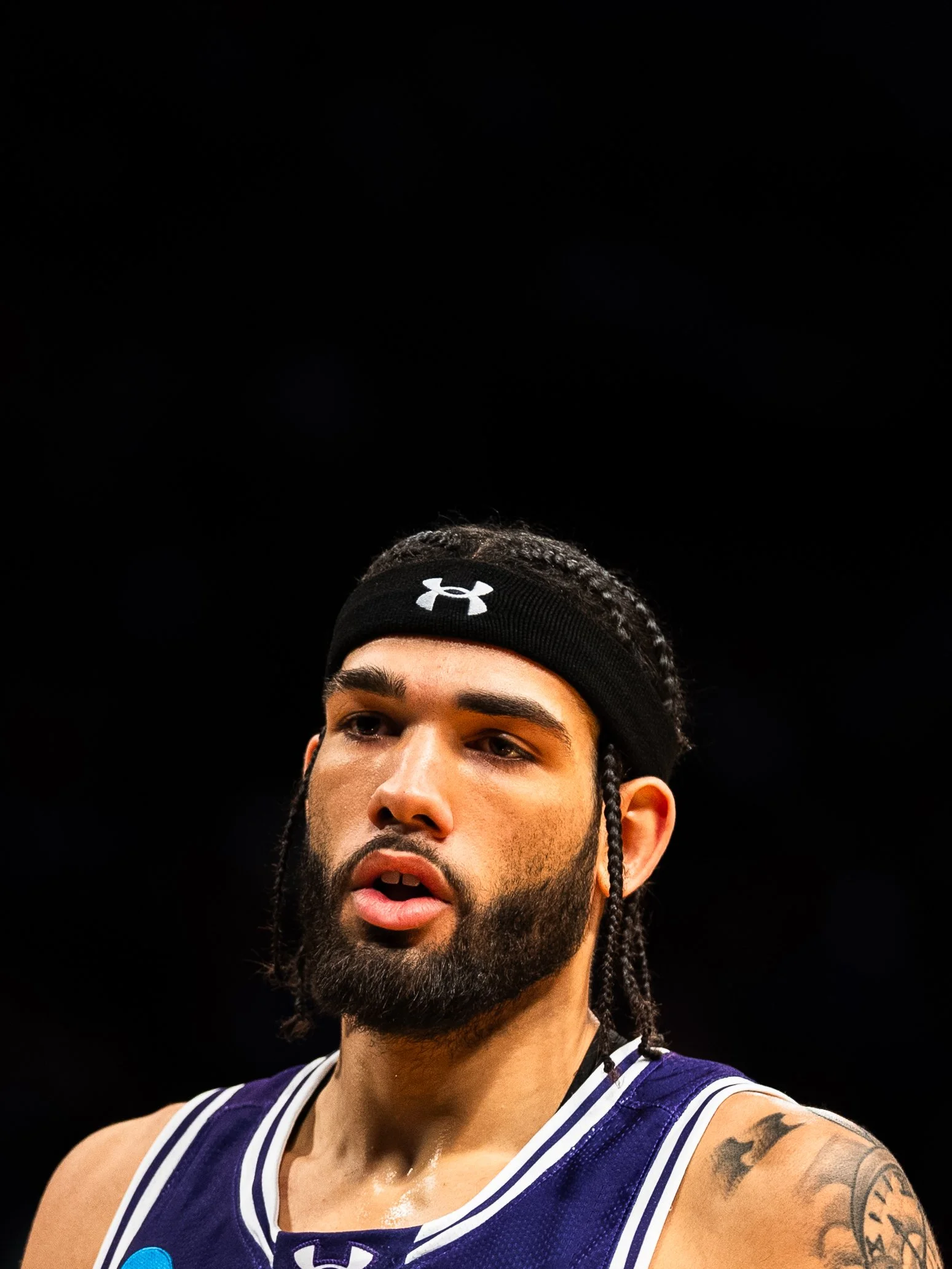 Close-up of a male basketball player with a beard and braided hair, wearing a black Under Armour headband and a blue jersey, against a dark background.