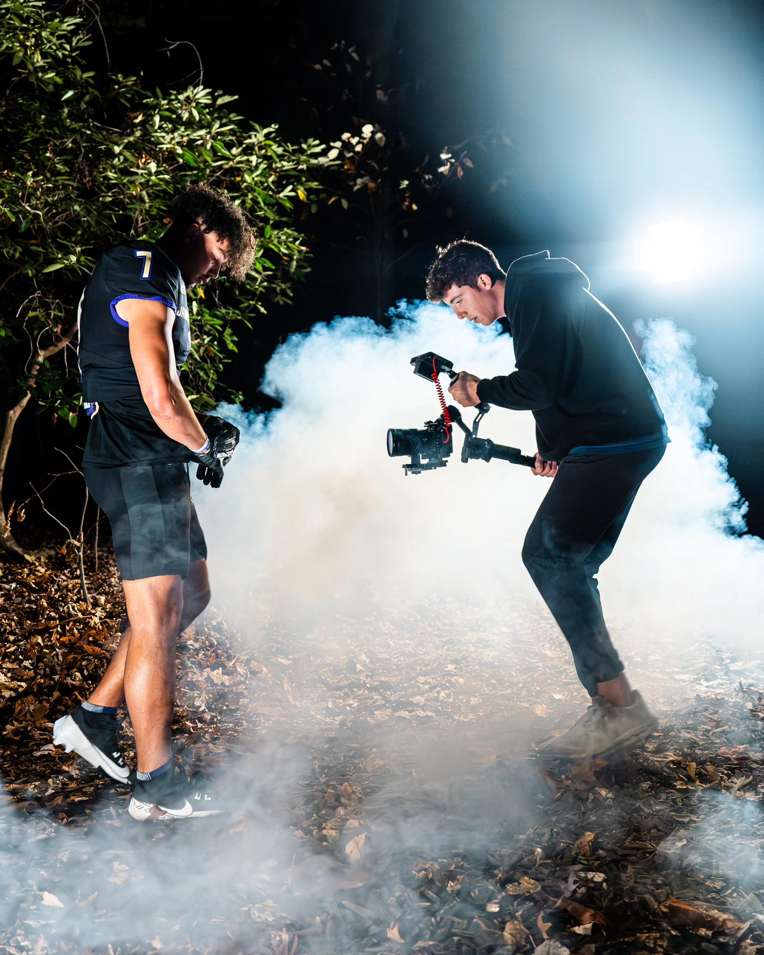 Two young men filming in a foggy forest at night, one with a camera stabilizer and the other in athletic clothing, surrounded by smoke and trees.