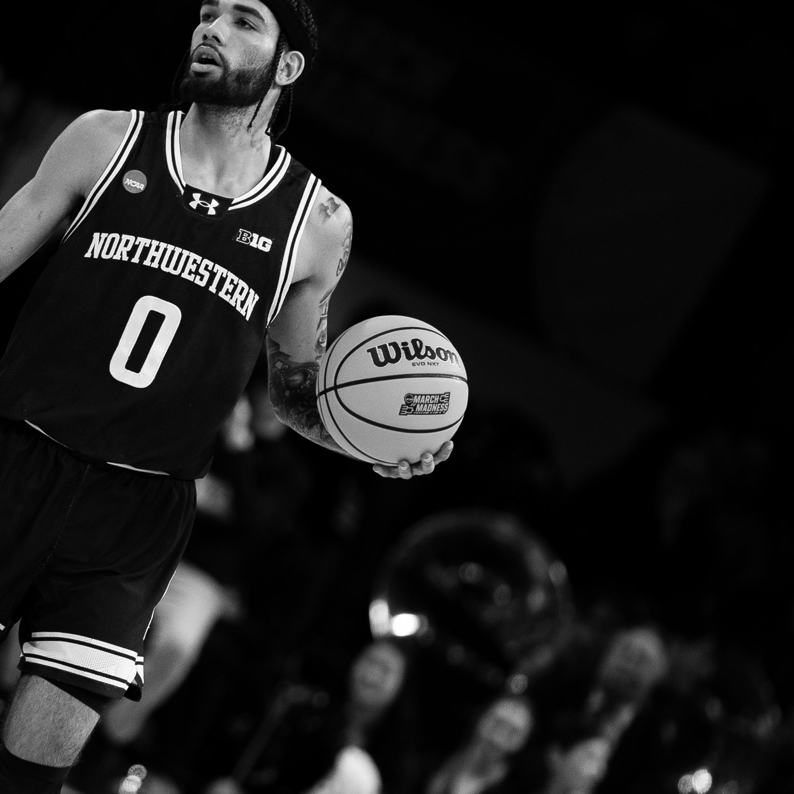 A basketball player with a beard and tattoos on his arms is holding a Wilson basketball on a basketball court. He is wearing a Northwestern basketball jersey with the number 0. The photo is in black and white.