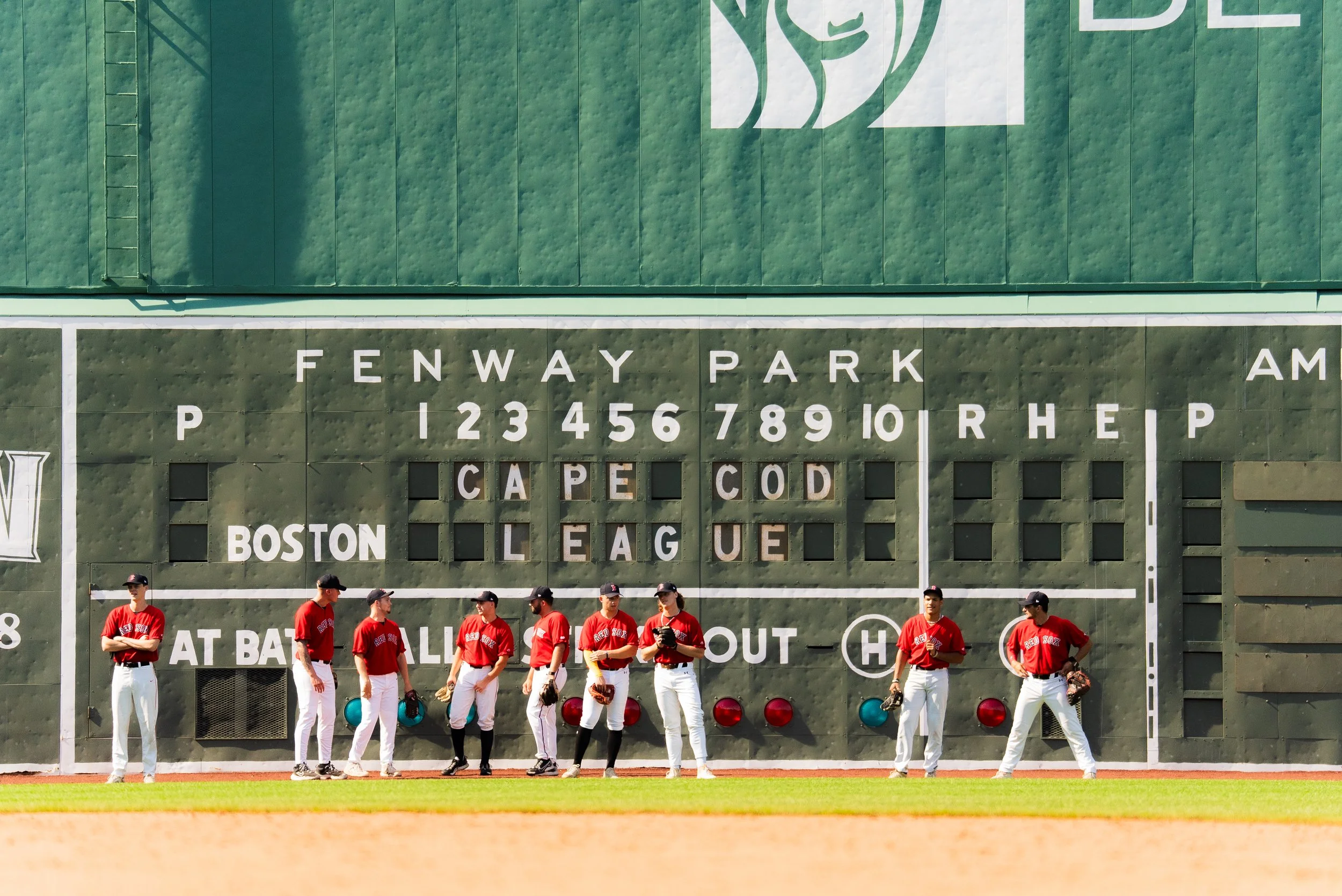 A group of baseball players standing in front of a green scoreboard at Fenway Park, Boston, wearing red jerseys and white pants.
