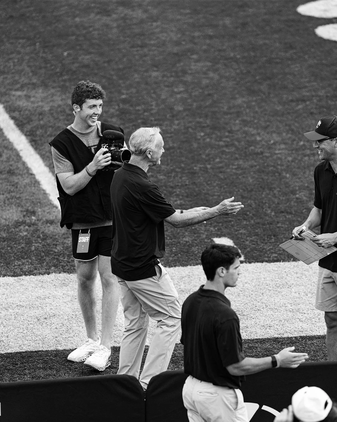 A group of people on a sports field, including a man with a camera, an elderly man reaching out, and a young man with a whistle, engaging in conversation.