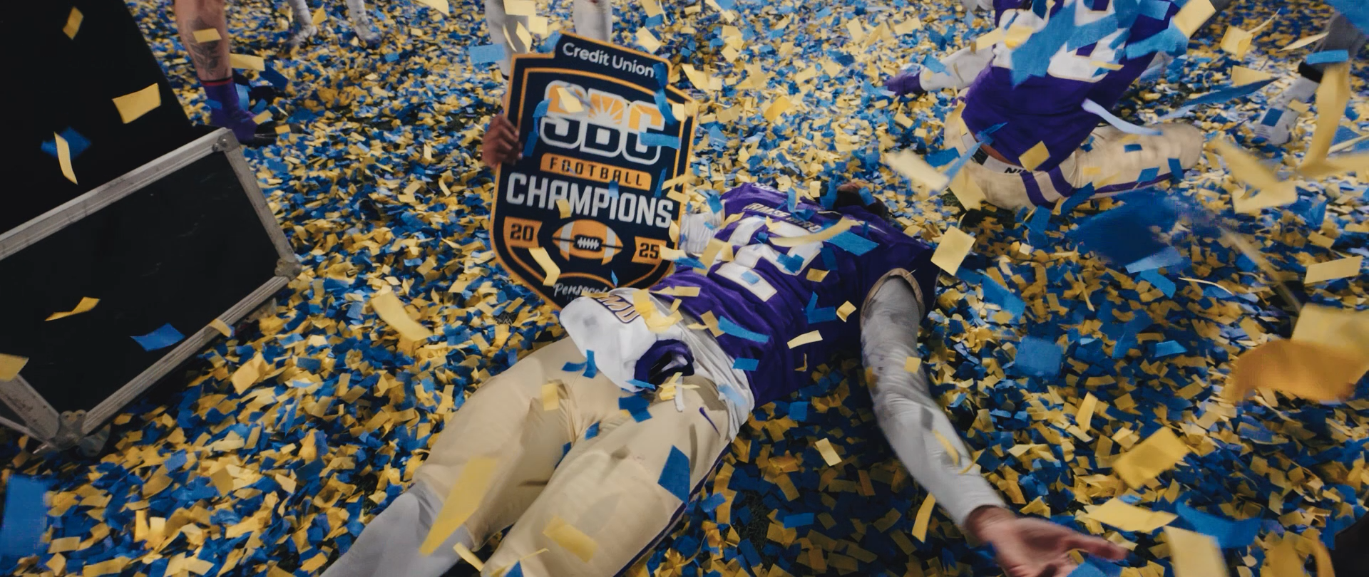 A football player in a purple uniform lying face down on a field covered in yellow and blue confetti. The player is holding a sign that says 'FCC Football Champions 2025' with a football graphic. Confetti is scattered across the ground and in the air, celebrating a victory.