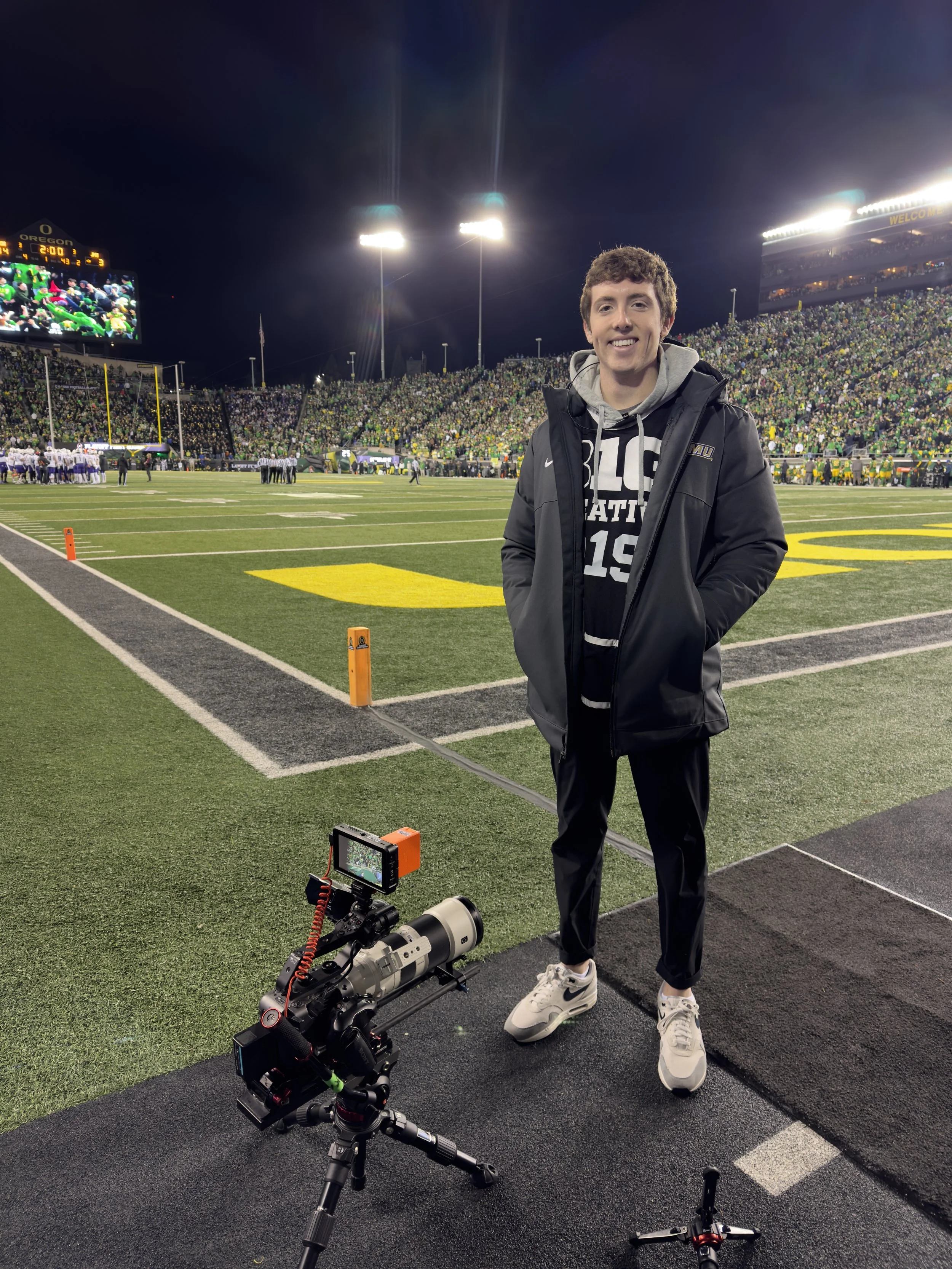 A young man standing on the sideline of a lit-up football stadium at night, with a camera on a tripod in front of him, and a large crowd in the stands.