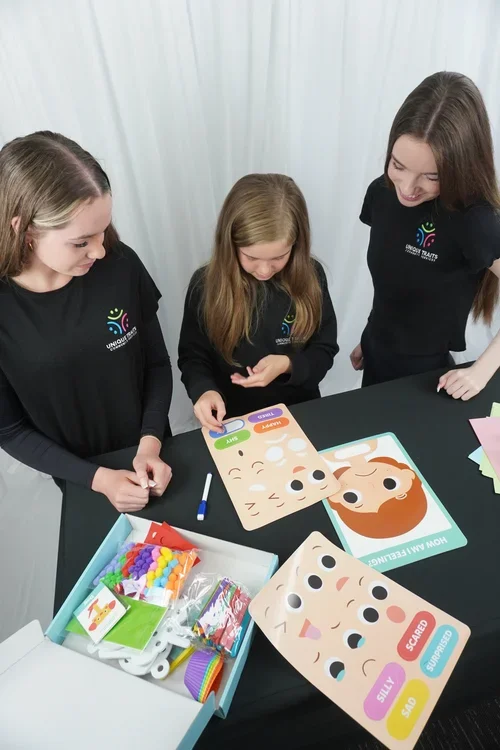 Three girls playing with face character boards and crafting supplies on a table.