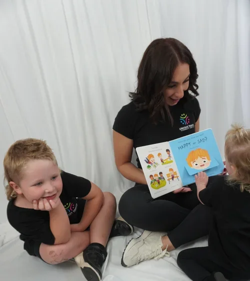 A woman reading a children's book titled 'HAPPY or SAD?' to two kids sitting on the floor against a white curtain background.