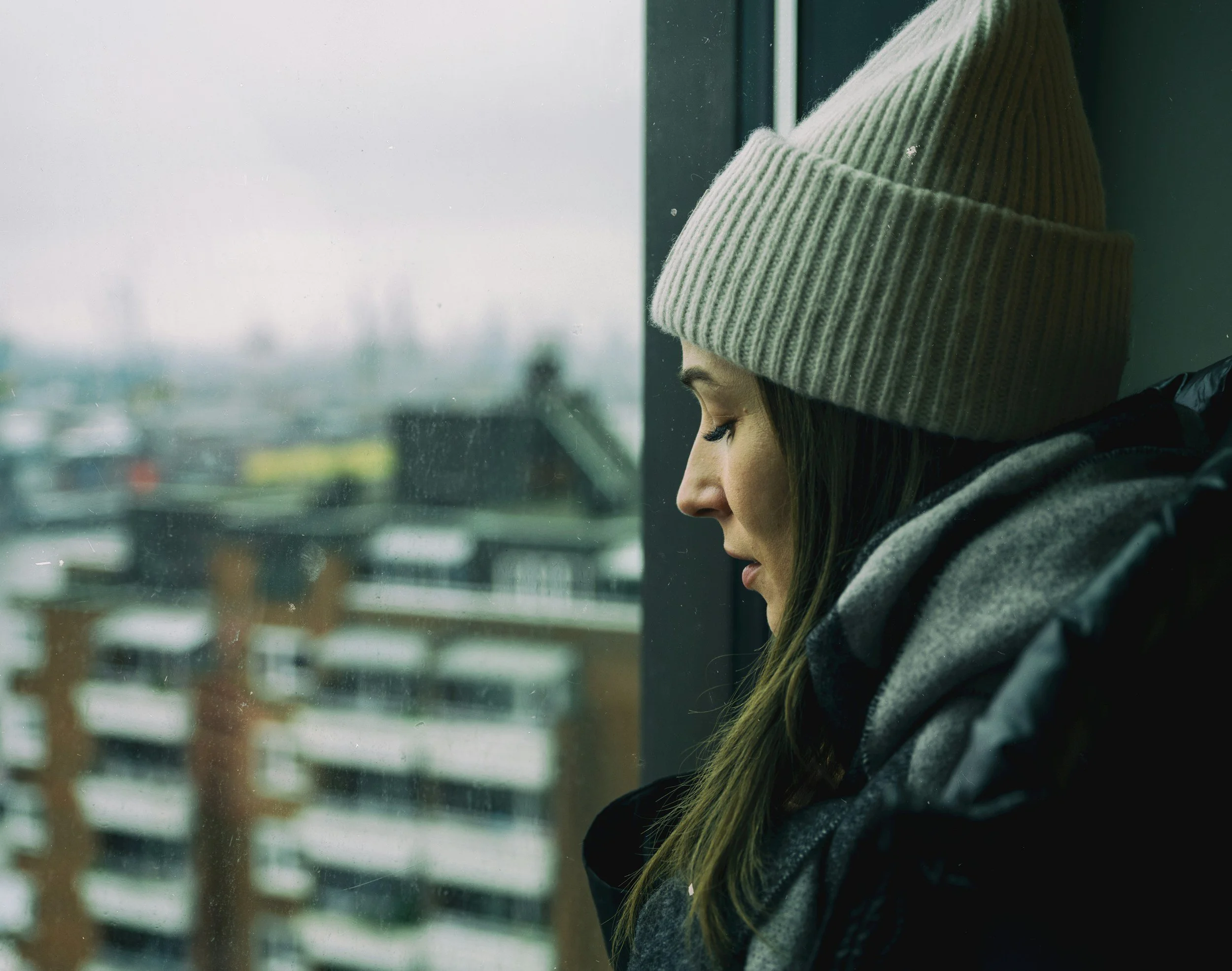 A woman wearing a beige knit beanie, gray scarf, and black winter coat sitting by a window with a view of snowy rooftops, looking down with a contemplative expression.