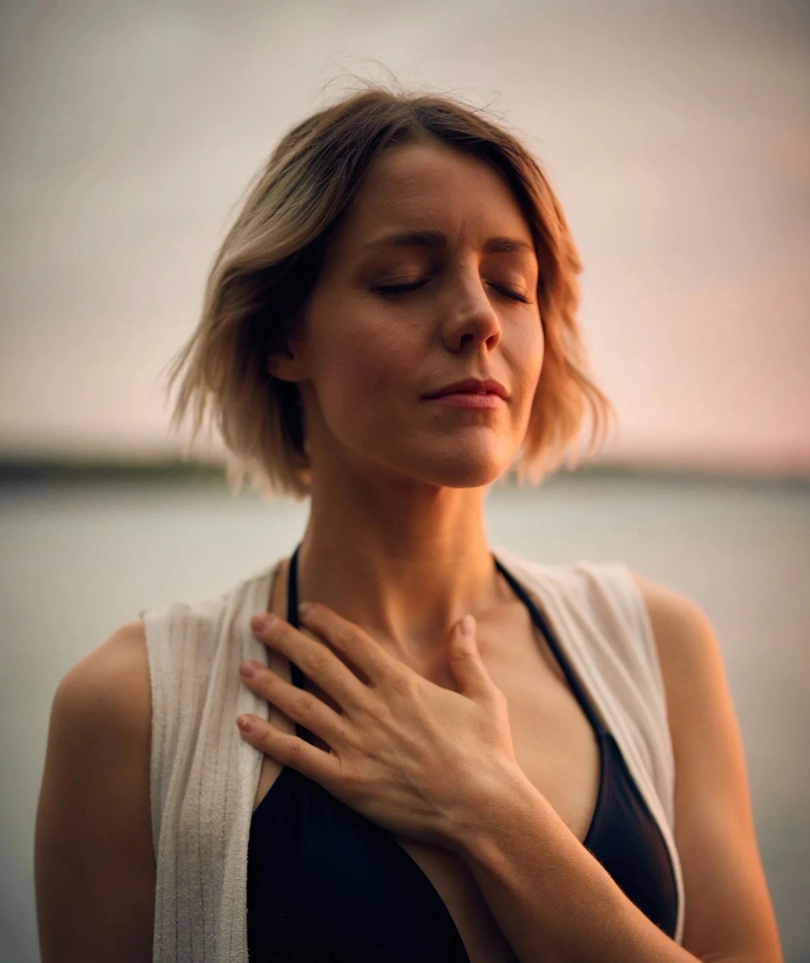 A woman with short blonde hair has her eyes closed and is touching her chest with one hand, outdoors at sunset.