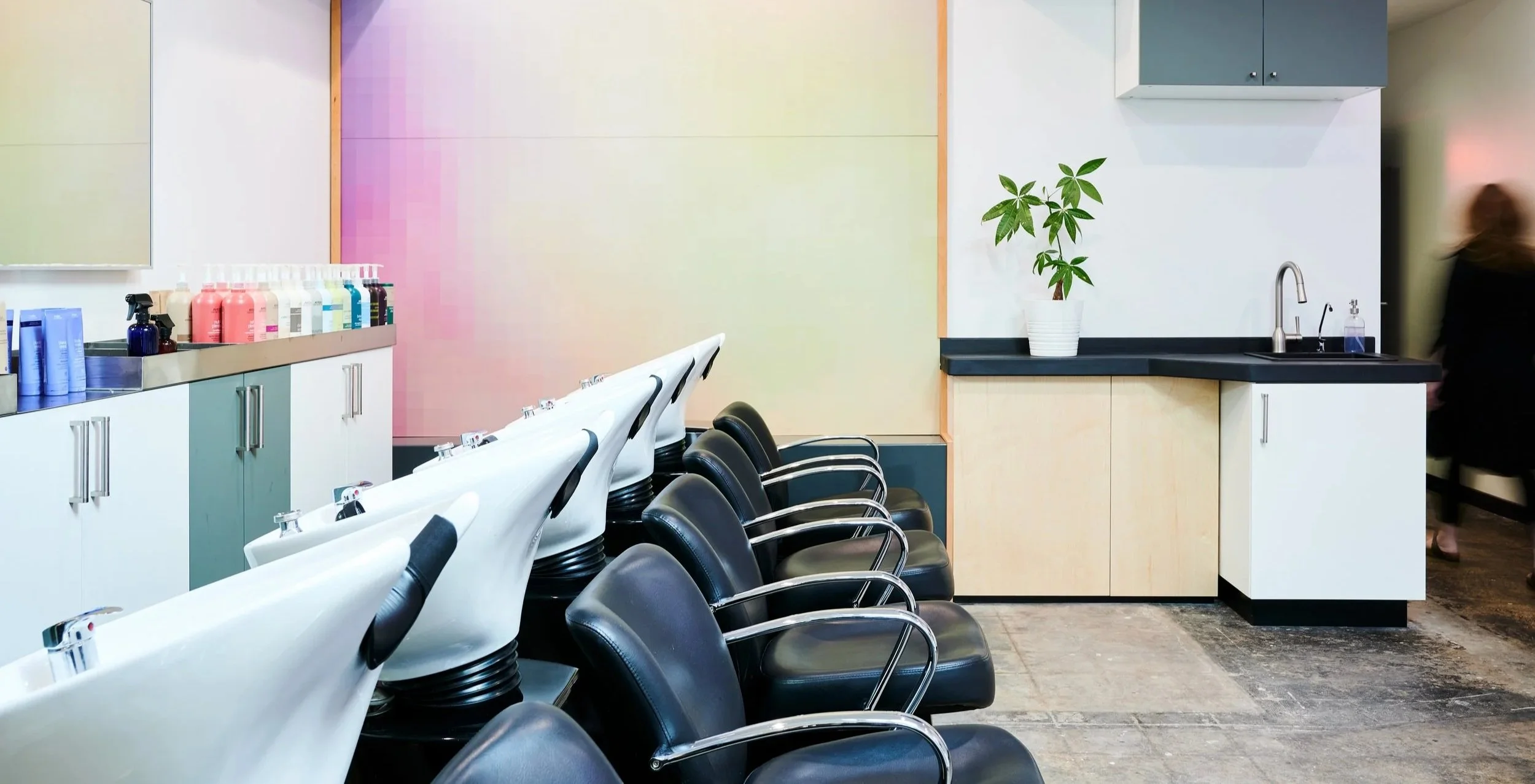 Salon shampoo bowls with black chairs, a sink, colorful hair product bottles, a plant on a black countertop, and a blurred person walking in the background.