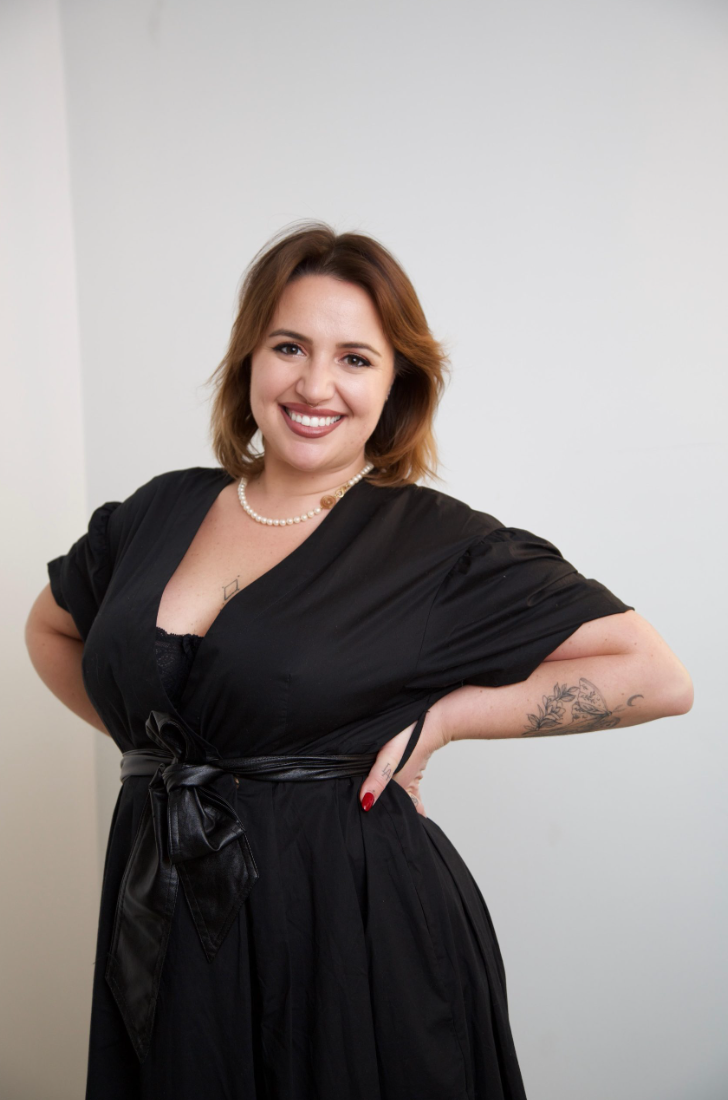 A smiling woman with shoulder-length auburn hair wearing a black dress with a bow at the waist, pearl necklace, and tattoos on her left arm poses with her hands on her hips against a plain white background.