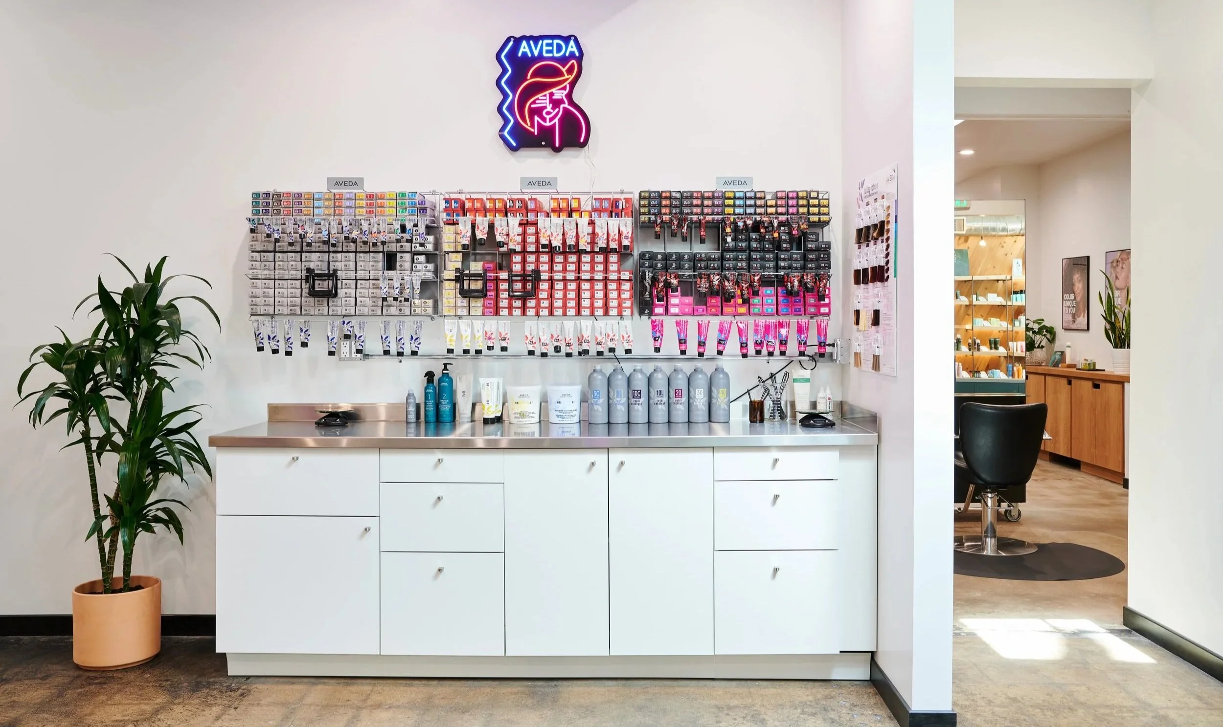 Display shelf with hair care products and tools, neon sign reading 'AVEDA' on the wall, potted plant to the left, and a seating area to the right, inside a modern salon.
