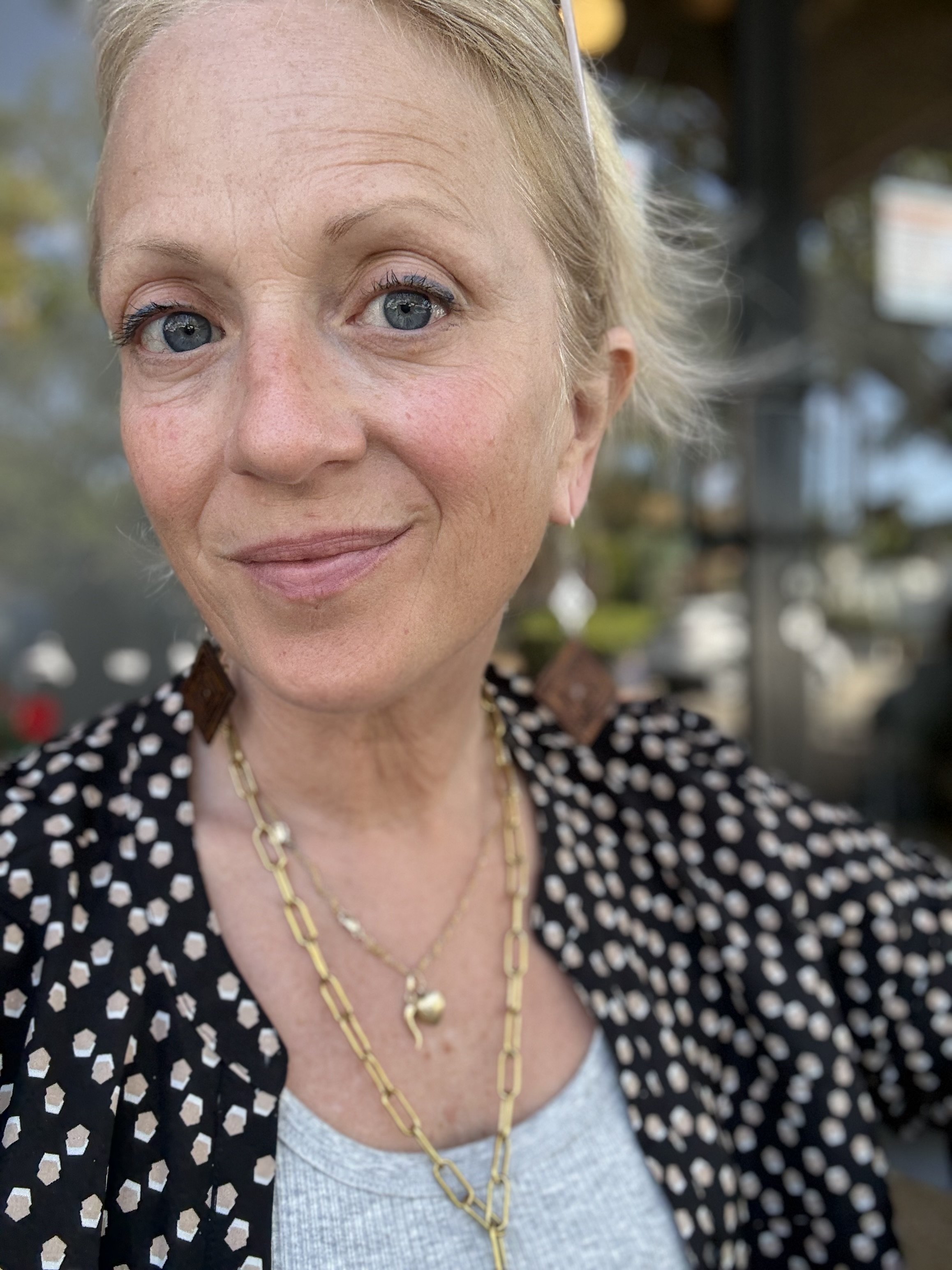 Close-up of a woman with lblue eyes, wearing a black and white polka dot blazer, a white top, and layered gold necklaces, outdoors with blurred background.
