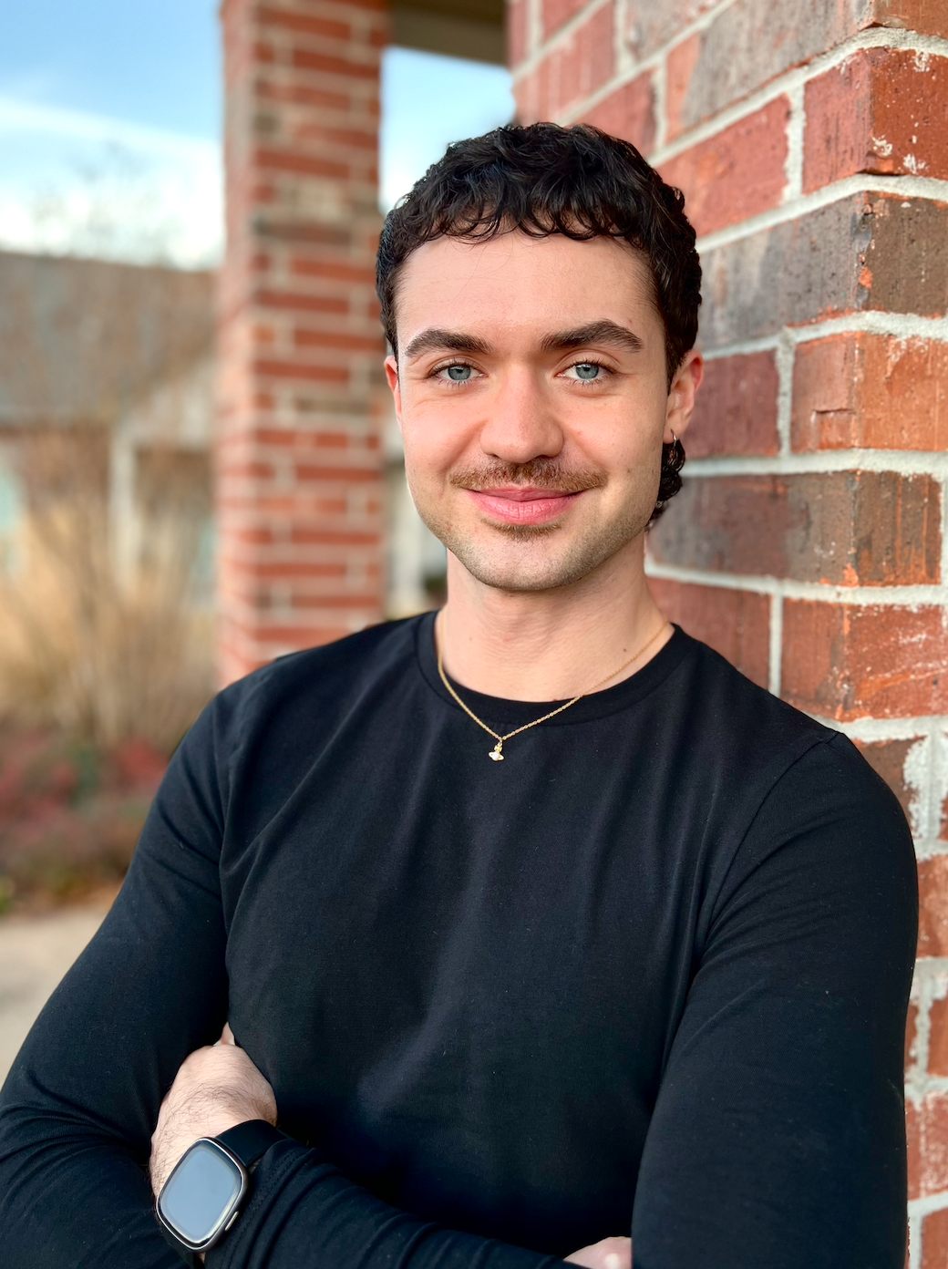 Young man with dark curly hair and blue eyes smiling, wearing a black shirt, gold chain necklace, and smartwatch, standing against a brick wall outdoors.