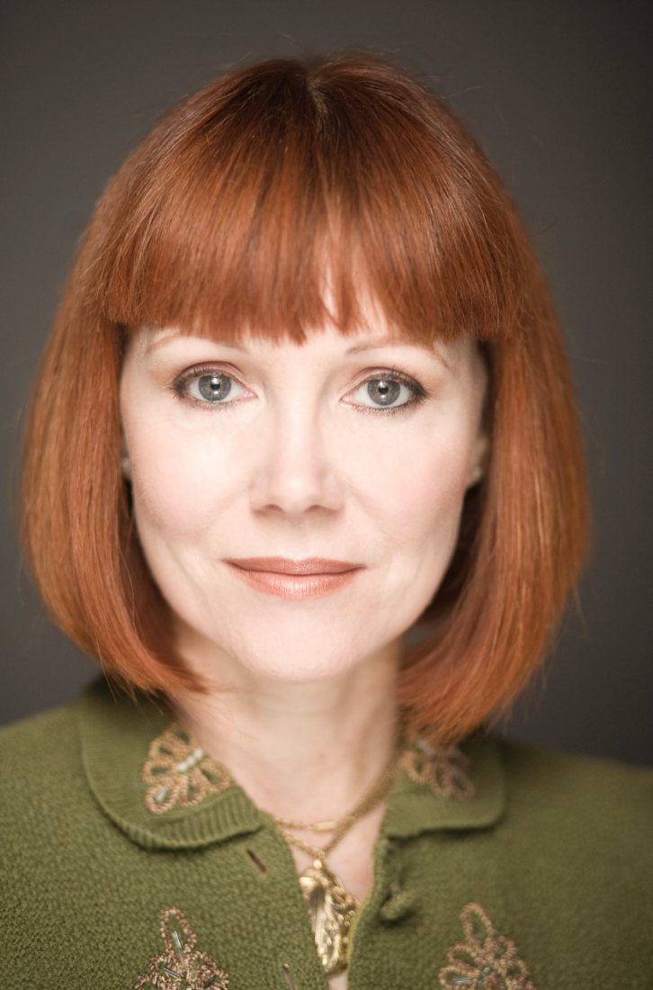 Close-up portrait of a woman with shoulder-length red hair, wearing a green top with embroidered details, gold jewelry, and light makeup, smiling gently against a dark background.